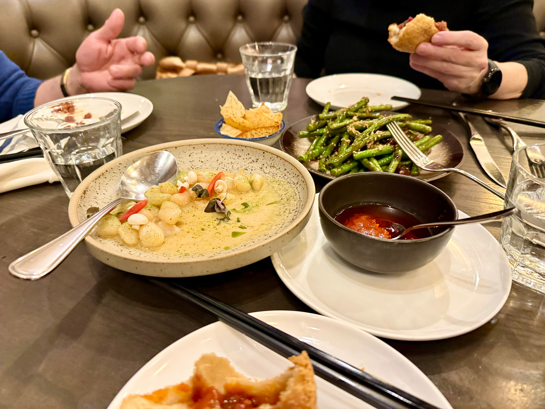 A table setting with various dishes, such as a creamy gnocchi in a speckled bowl garnished with thinly sliced peppers and herbs. Next to it is a plate of sautéed green beans with sesame seeds, a small bowl of red sauce with a spoon, and a plate of crispy crackers. Two people are seated, one holding a piece of bread, with glasses of water and cutlery visible on the table.