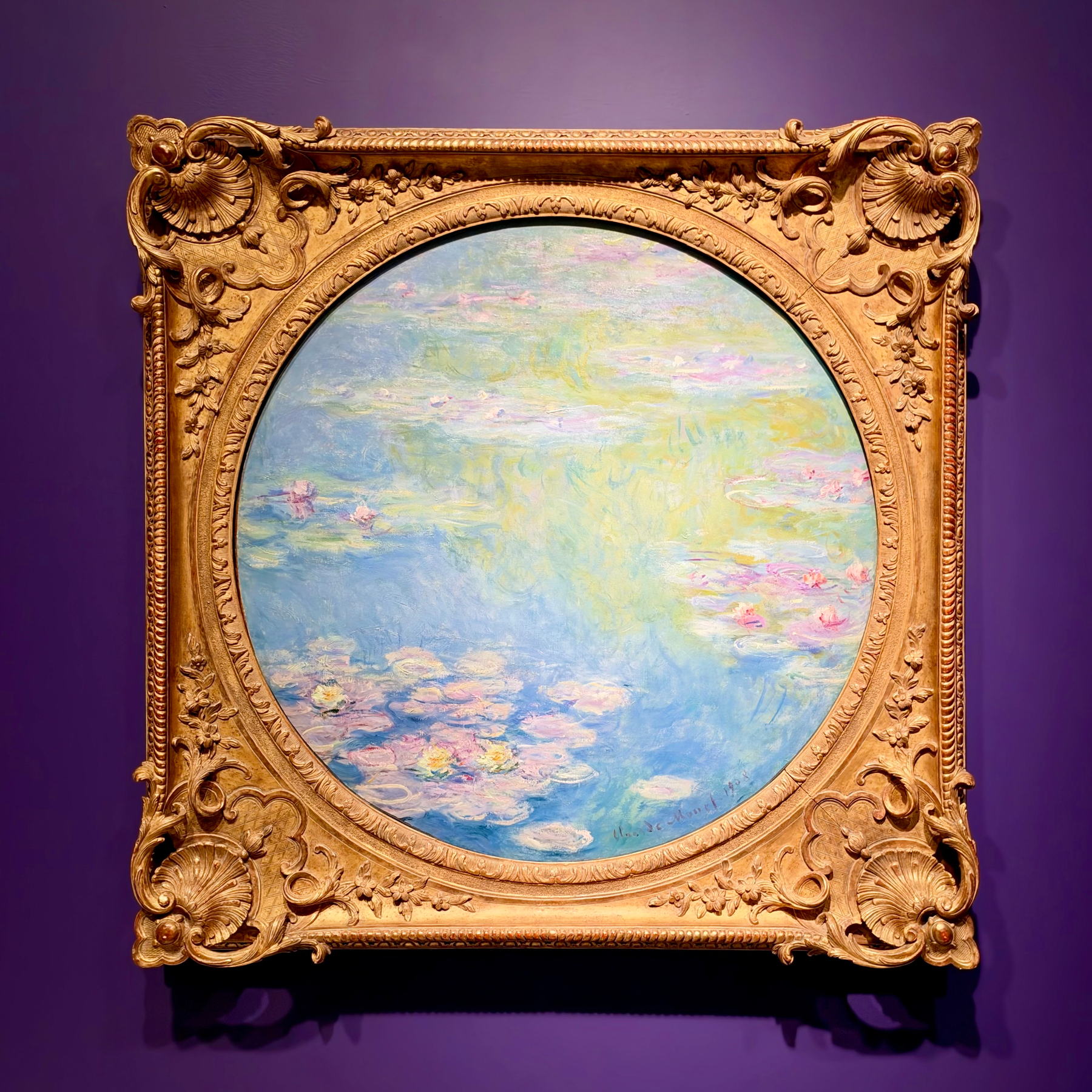 Water Lilies (Nymphéas) by Claude Monet, 1908&10;A circular painting in an ornate, gold, baroque-style frame hangs on a deep purple wall. The painting depicts a serene water scene with soft, impressionistic brushstrokes in pastel hues of blue, green, pink, and lavender. Water lilies float across the surface, primarily in the lower portion, blending harmoniously with the rippling reflections of sky and foliage. The thick, decorative frame features carved seashell motifs, scrolling patterns, and floral accents at the corners and along the edges. The artwork is illuminated to highlight its colours and textures against the smooth, dark background.