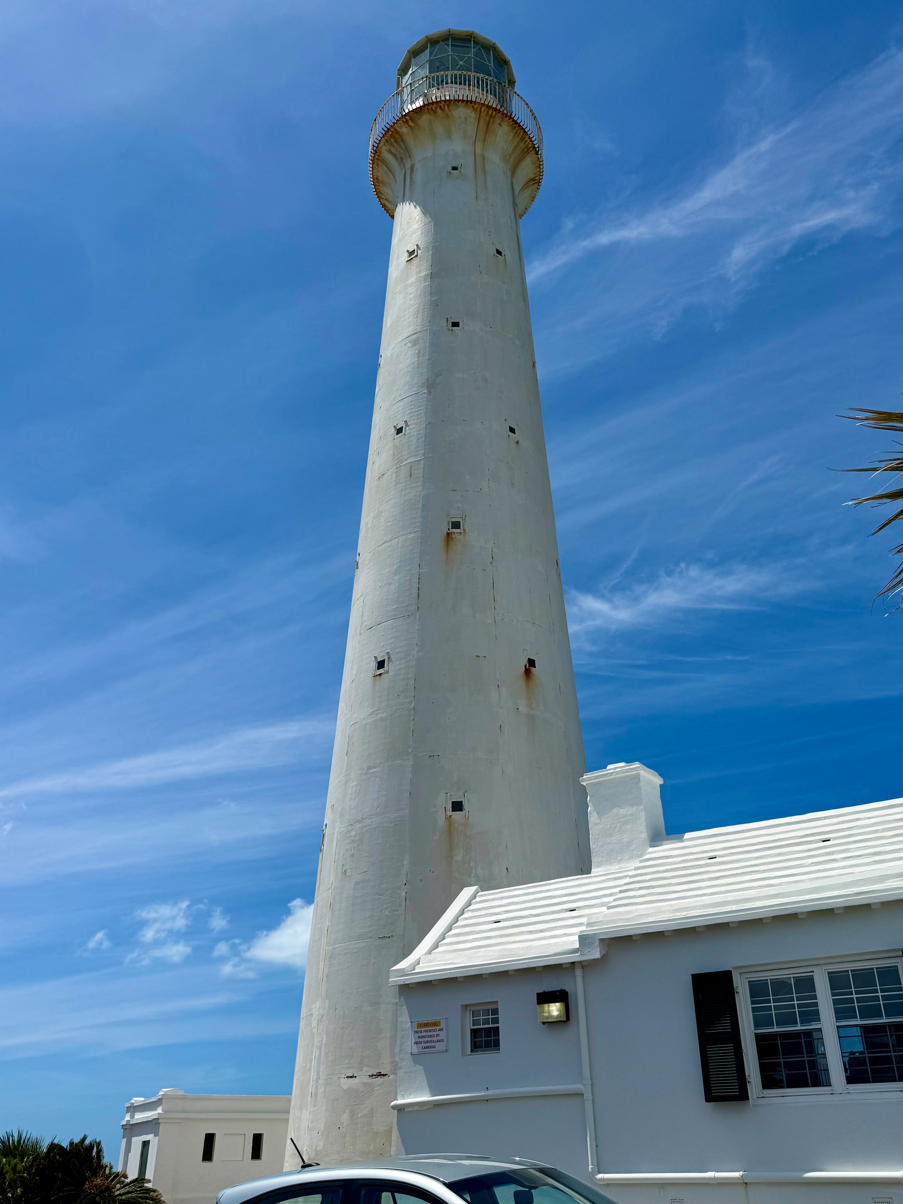 A tall, white lighthouse rises into a bright blue sky with wispy clouds. The cylindrical structure tapers as it reaches the top, where a circular observation platform with a rusted railing surrounds the lantern room. The lighthouse has several small rectangular windows spaced irregularly along its height, and there are patches of weathering and rust near some edges. Attached to the base is a white building with a sloped roof and black shutters on its windows. A silver car is parked in front of the building, and palm fronds are partially visible along the bottom edge of the image.