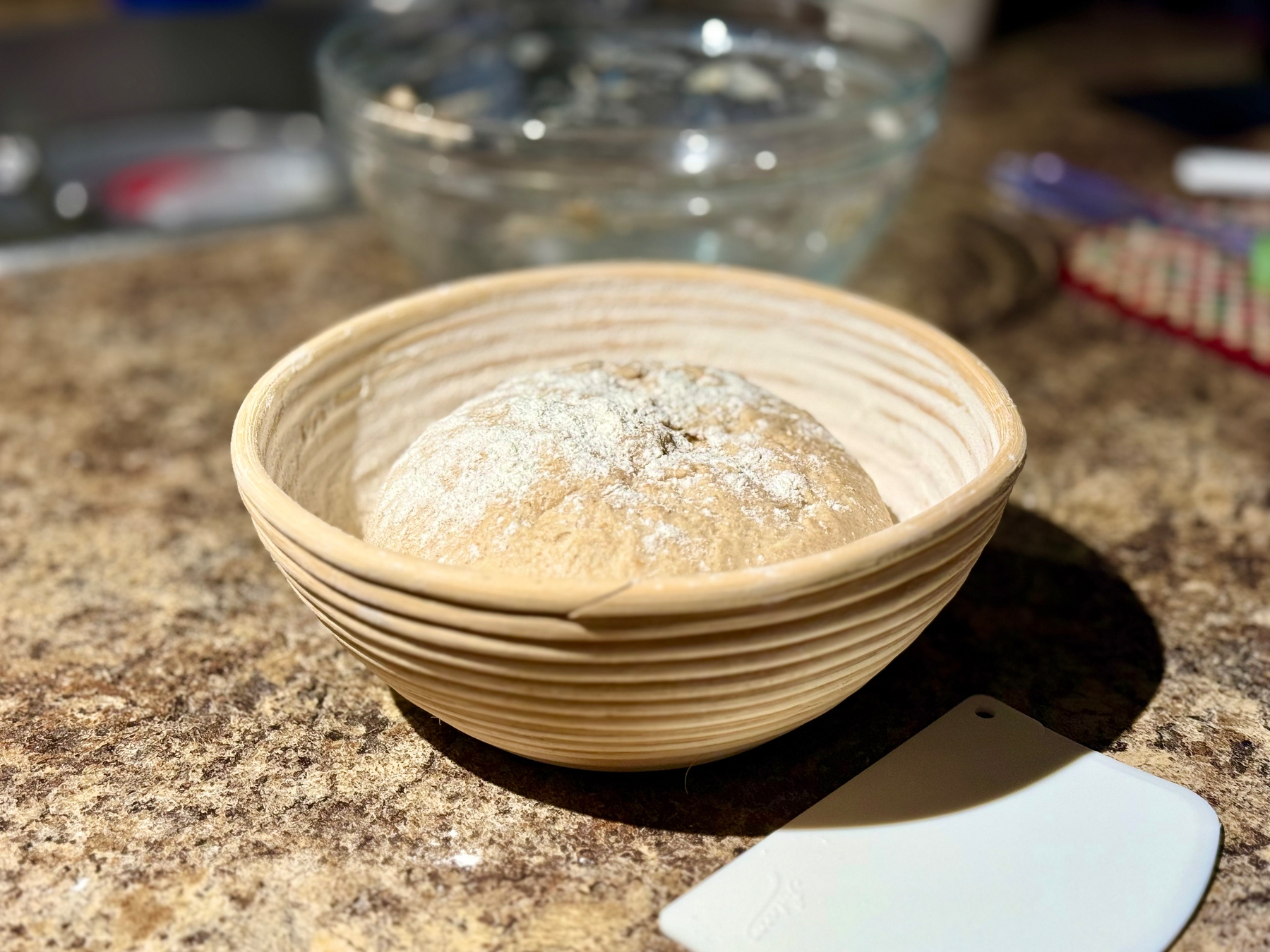 A round dough is resting in a wooden banneton basket on a kitchen countertop. The dough is lightly dusted with flour. In the background, there is a glass bowl, and a plastic dough scraper is placed nearby. The lighting highlights the texture and color of the countertop and the dough.
