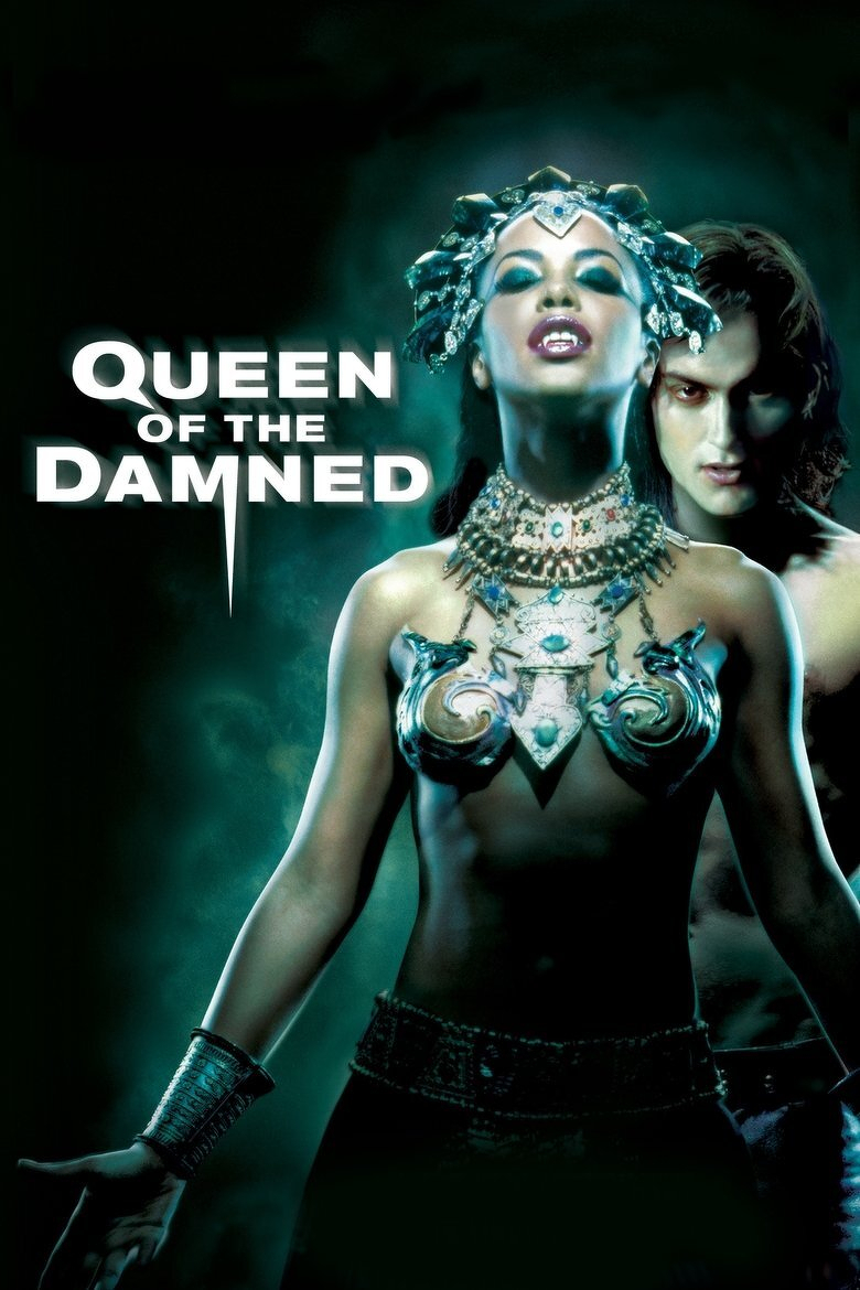 A dark, moody poster featuring a woman in elaborate, metallic, and jewel-encrusted Egyptian-inspired armor, including a headdress, necklace, and ornate bra shaped like animal figures. Her eyes are closed, head tilted back slightly, and she bares sharp fangs in a vampiric expression. Behind her, a pale man with long dark hair and an intense gaze lurks in the shadows, partially obscured. The background is a smoky, greenish-black mist. The white text on the left side of the image reads “QUEEN OF THE DAMNED,” with the word “OF” smaller and positioned between the larger words, and a sharp fang-like design extending downward from the letter "M."