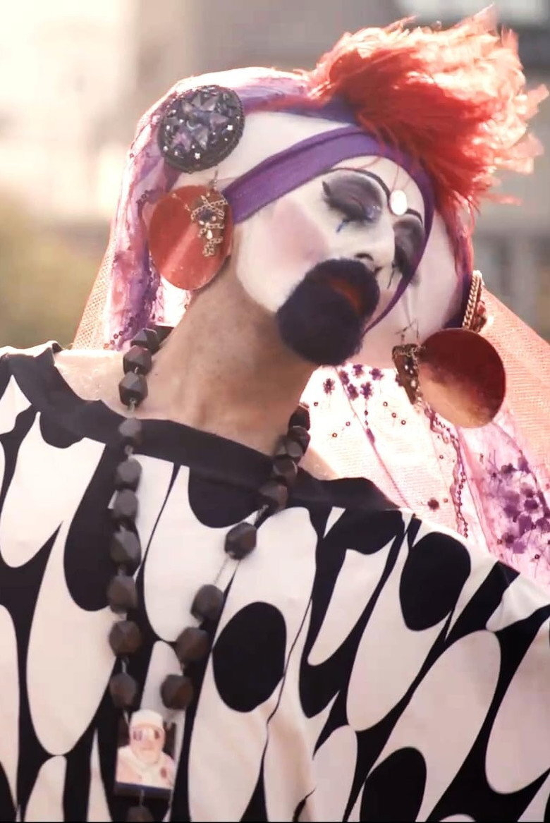 A person in elaborate makeup and costume poses with their head tilted. They are wearing white face paint with dramatic black and purple eye makeup, dark lipstick, and a painted black beard. A small jewel is placed on their forehead. Their head is adorned with a bright red feather-like headpiece, a purple band, and a pink mesh veil decorated with purple flowers and sequins. Large circular red earrings with dangling charms and a big black gemstone brooch are attached to the headpiece. They wear a black-and-white patterned outfit with large abstract shapes and a chunky black beaded necklace featuring a small charm showing a similar costumed figure. The background is softly blurred, suggesting an outdoor setting.