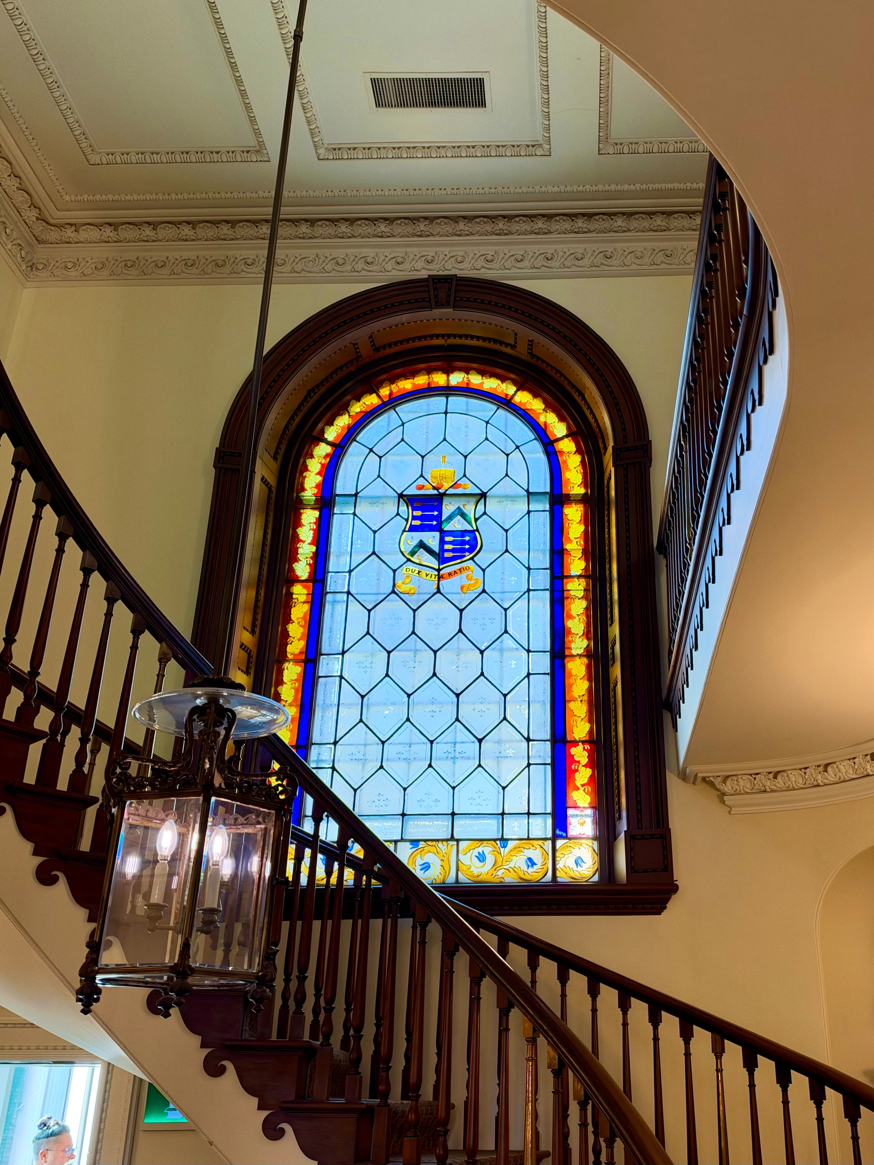 A grand interior staircase with dark wooden railings curves around a large, arched stained-glass window. The window features a geometric pattern of hexagonal glass panes with a colourful border and a central design of a blue and gold heraldic shield topped with a gold crown. The shield has a blue and white pattern with a gold ribbon beneath. The outer stained-glass border alternates between warm golden tones and deep blue accents, with a row of yellow floral motifs at the bottom. A vintage hanging lantern with multiple lit bulbs hangs above the staircase, and the ceiling is adorned with ornate molding and a recessed vent. A small portion of a person’s head is visible at the bottom left near another window.