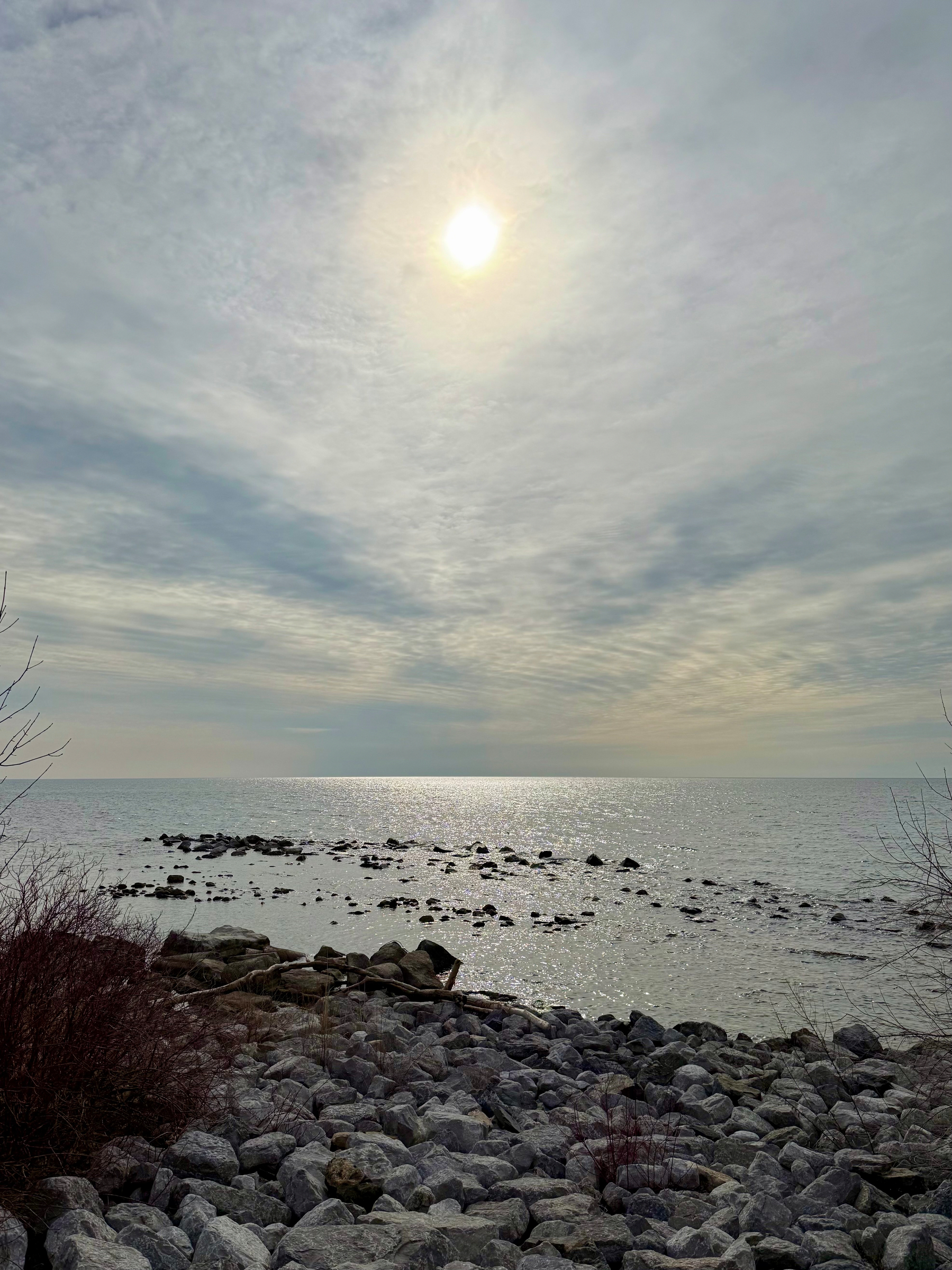 A rocky shoreline stretches into a calm body of water under an overcast sky. The sun is partially obscured by thin clouds, casting a soft, diffused light that reflects off the gentle ripples on the water’s surface. In the foreground, large grey rocks and sparse, leafless shrubs line the shore. Further out, small clusters of rocks emerge from the water, creating a natural pattern across the horizon. The overall scene conveys a quiet, serene atmosphere.