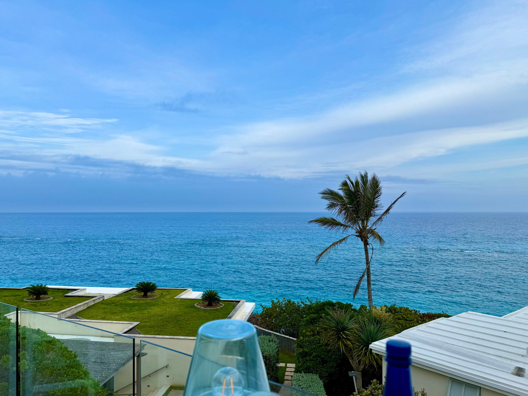 A scenic view of the ocean under a partly cloudy blue sky, with shades of turquoise and deep blue water stretching to the horizon. In the foreground, a glass balcony overlooks a modern building with a grassy rooftop and small round planters holding palm-like plants. A single tall palm tree rises near the shoreline, swaying slightly toward the ocean. Lush green vegetation surrounds the buildings, and a white-roofed structure is partially visible on the right. A transparent glass and the top of a blue bottle sit on the balcony railing, slightly out of focus.