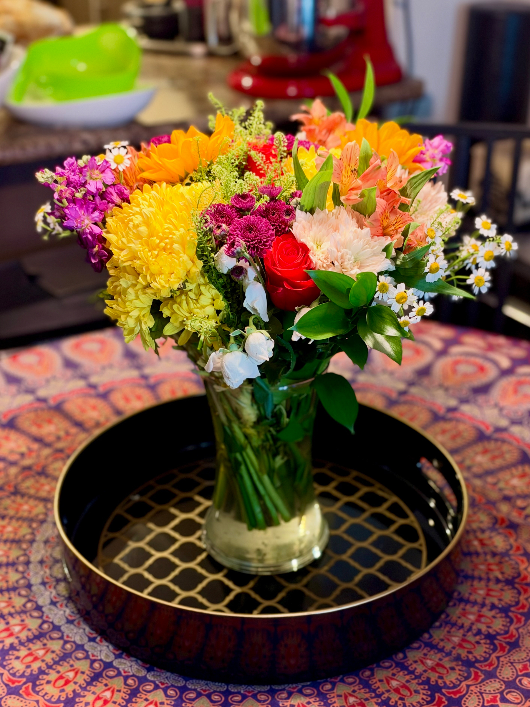 A vibrant bouquet of flowers in a glass vase sits on a patterned round tray placed on a table with a colourful, intricate tablecloth featuring red, purple, and pink designs. The bouquet includes a mix of yellow chrysanthemums, orange lilies, white daisies with yellow centres, bright purple flowers, small magenta blooms, a single red rose, and lush green leaves. The stems are visible through the clear vase filled with water. In the background, a kitchen counter holds a green bowl, a white bowl, and a red stand mixer, with other kitchen items slightly blurred.
