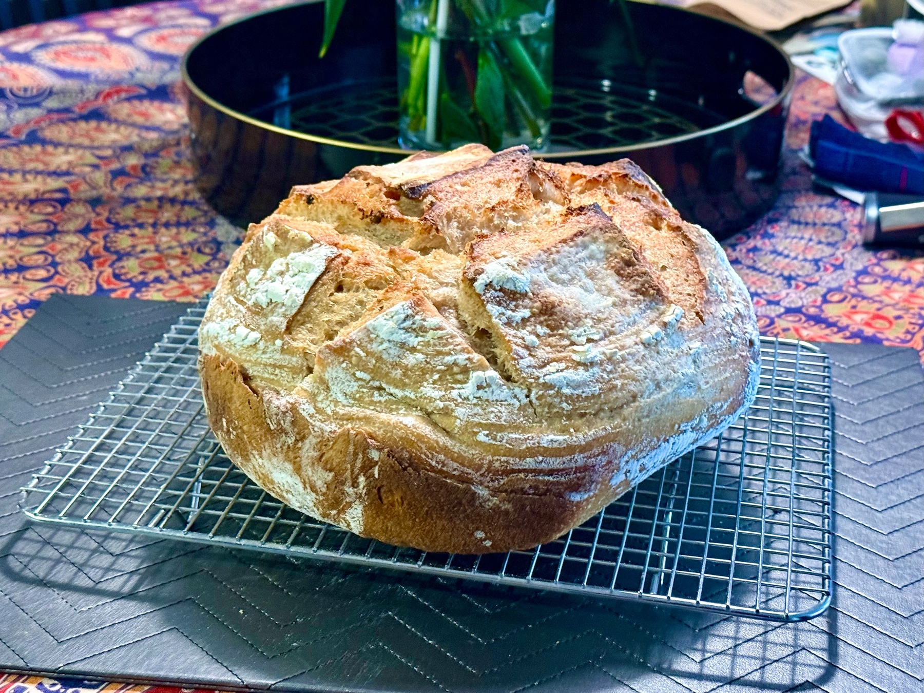 A freshly baked round loaf of rustic bread with a golden-brown crust and light dusting of flour sits on a metal cooling rack. The bread has deep, irregular scoring patterns that have opened up during baking, creating a textured surface. The cooling rack rests on a dark, quilted-pattern mat on a colourful patterned tablecloth. In the background, there is a black circular tray with a vase of green leaves, and various items like folded fabric and containers are partially visible.