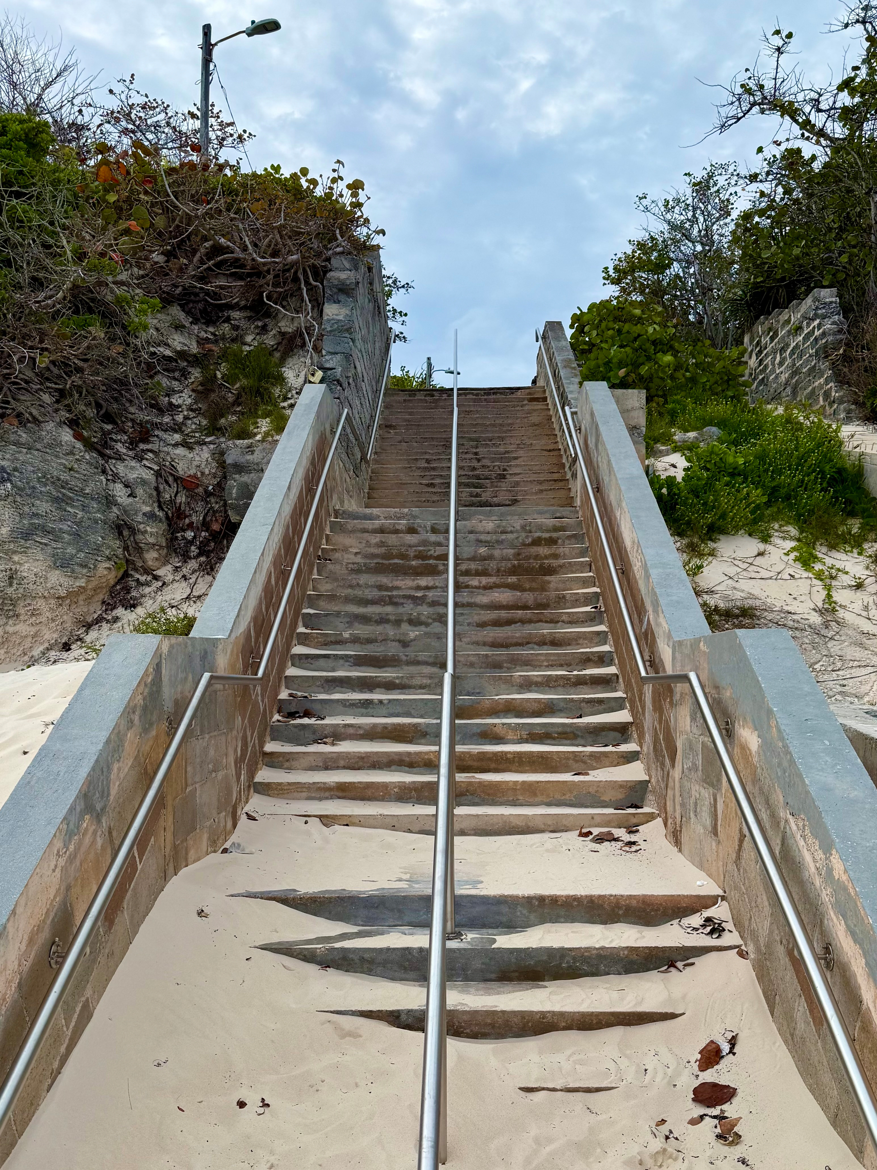 A steep outdoor stone staircase leads upward between two rocky embankments covered with green vegetation and shrubs. The steps are partially buried under windblown sand, especially at the lower section, where several steps are almost completely covered. A metal handrail runs up the centre of the staircase, with additional handrails along both sides attached to the stone walls. The sky above is overcast with patches of light blue visible, and a single streetlamp stands at the top left of the stairs.