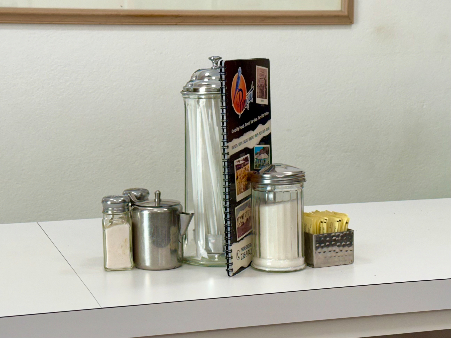 A white tabletop holds a small restaurant setup including a glass salt shaker, a small metal container with a spout, a tall glass sugar dispenser with a metal lid, a spiral-bound menu standing upright, a shorter glass sugar dispenser with a metal lid, and a metal holder with yellow sugar packets. The background is a light beige wall with a wooden-framed picture partially visible at the top.
