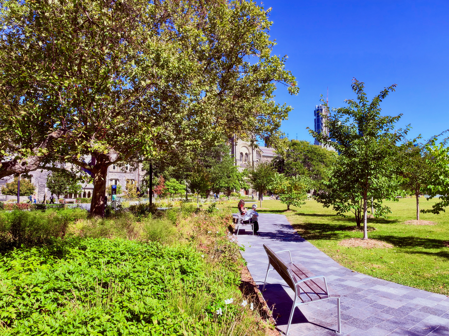 A sunny park scene with a paved walkway winding through lush green grass and trees. Two wooden benches with metal frames are along the path, one in the foreground and another occupied by two people further down the path. The park is filled with a variety of trees, some casting shade, and low green shrubs and plants on either side of the walkway. In the background, there are stone buildings with arched windows partially visible behind the trees, and a modern glass skyscraper rises further in the distance against a clear, bright blue sky. A few people are walking along a path near the stone building.