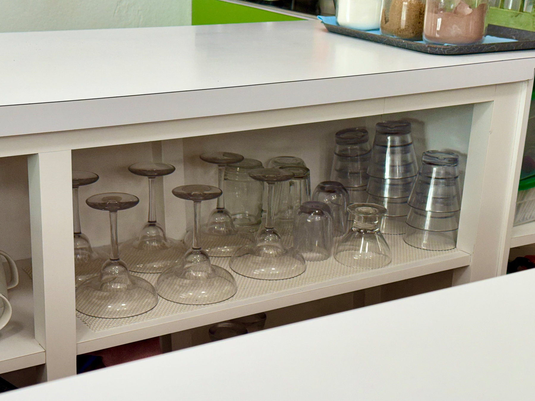 A white kitchen shelf with a collection of glassware neatly arranged. On the top shelf, there are several wine glasses placed upside down on a textured mat, along with some smaller tumblers and glasses also inverted. To the right, multiple plastic cups are stacked in two neat columns, all facing downwards. The surface above the shelf is bare except for a tray in the background holding jars filled with white sugar, brown sugar, and cocoa powder. The surrounding area includes hints of a green wall section and adjacent shelving.
