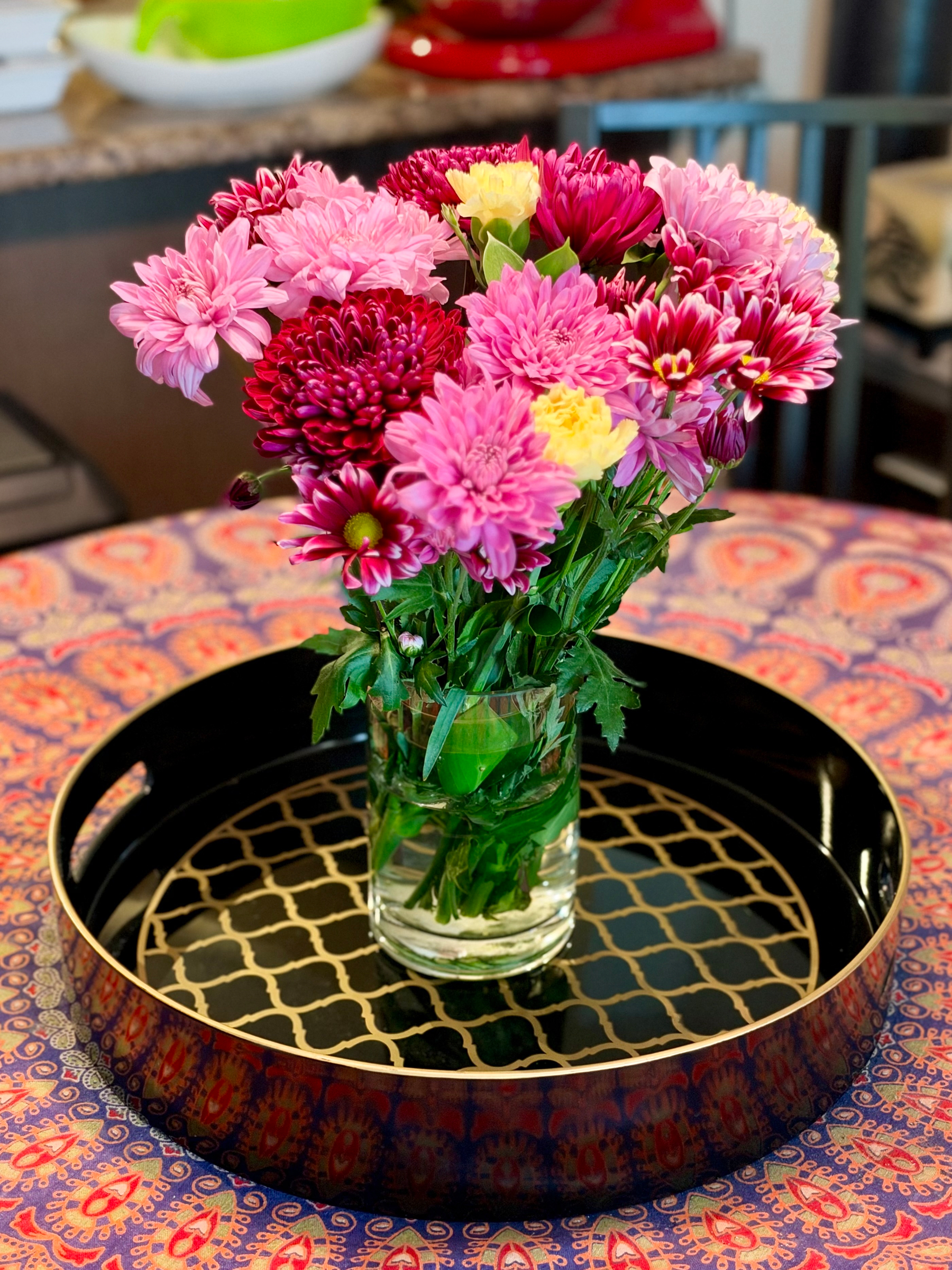 A clear glass cylinder vase holds a full bouquet of mostly chrysanthemums in pink, magenta, and deep burgundy, with a few pale yellow carnations tucked among them. Some blooms are shaggy and ruffled, others are daisy-like with white-tipped petals, and a few closed buds show along the stems. Green leaves and stems are visible through the water. The vase sits on a black tray with a gold rim, handles on each side, and a gold quatrefoil lattice pattern across the base. The tray rests on a round table covered in a blue, orange, and pink paisley-medallion tablecloth. Behind the arrangement, a blurred kitchen shows a granite countertop, a white bowl with a green item, a red appliance, and a dark metal chair. No words are visible in the photo.