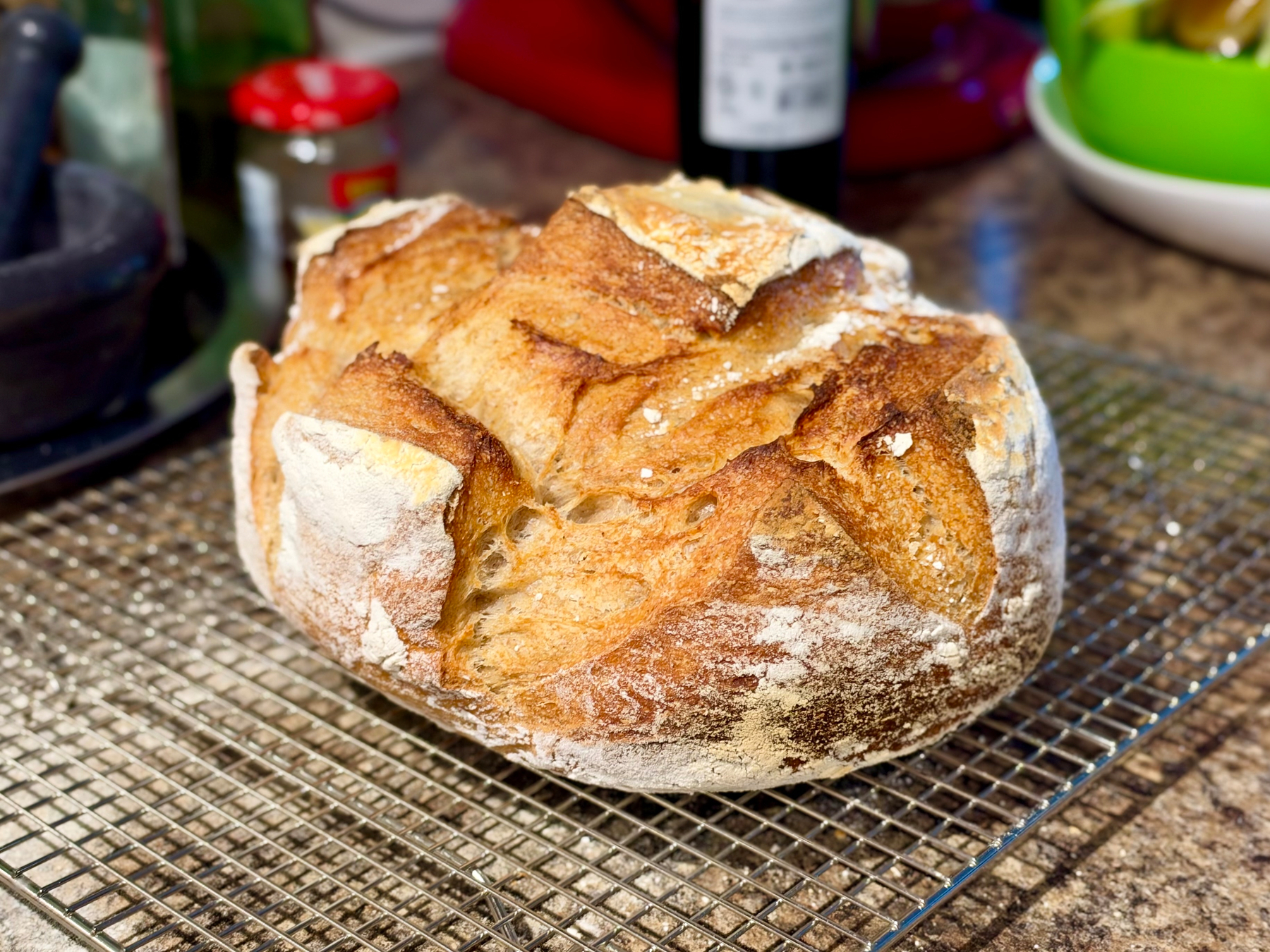 A round, rustic artisan loaf sits on a silver wire cooling rack on a speckled granite countertop. The crust is deep golden brown with darker, caramelized patches, and its surface is dusted with flour. The top is slashed with irregular, overlapping cuts that opened into pale, ridged flaps, and a small opening in one score reveals the airy, open crumb inside. In the blurred background are kitchen items: a dark mortar and pestle on the left, a glass jar with a red lid, a dark wine bottle, a red appliance, and a green and white bowl in the upper right corner.