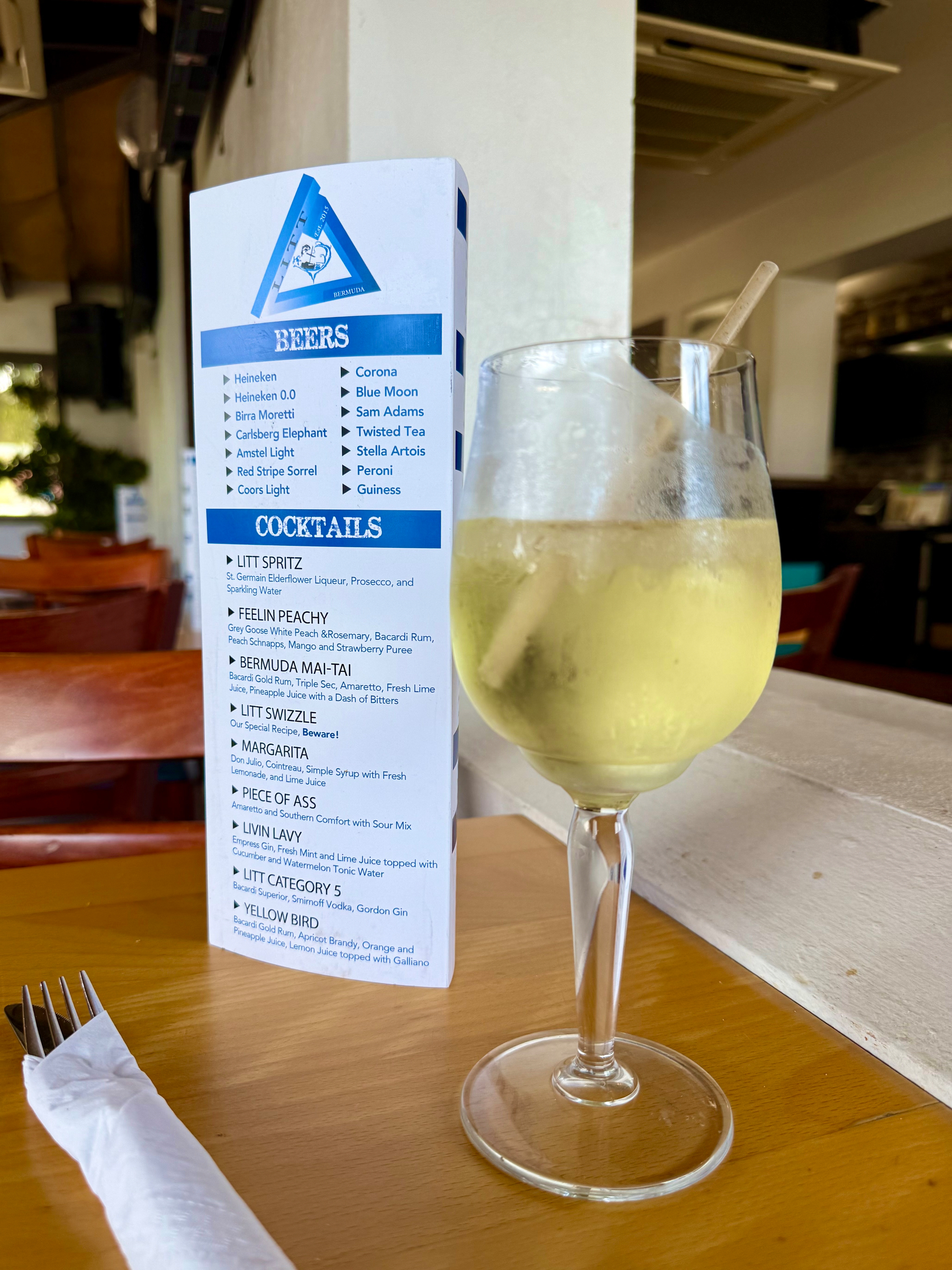 A glass of chilled white wine with a straw sits on a wooden table in a restaurant. Beside it is a folded drink menu showing a list of beers and cocktails. The beers section includes options like Heineken, Heineken 0.0, Bira Moretti, Carlsberg Elephant, Amstel Light, Red Stripe Sorrel, Coors Light, Corona, Blue Moon, Sam Adams, Twisted Tea, Stella Artois, Peroni, and Guinness. The cocktails section lists drinks such as Litt Spritz, Feelin Peachy, Bermuda Mai-Tai, Litt Swizzle, Margarita, Piece of Ass, Livin Lavy, Litt Category 5, and Yellow Bird, with ingredients described under each. A fork wrapped in a napkin lies on the table. In the background, there are wooden chairs, part of a white partition wall, and a mix of indoor and outdoor seating with some greenery visible outside.