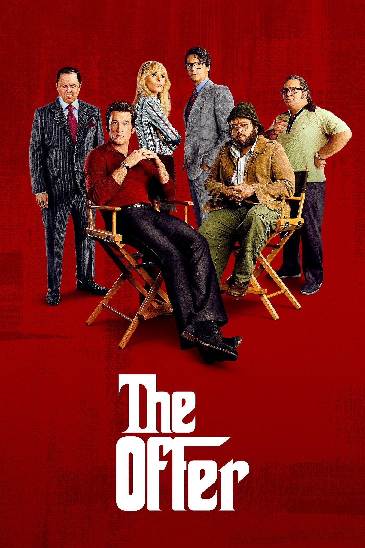 Six people standing and sitting against a solid red background. Two men are seated in wooden director’s chairs at the front. The man on the left wears a fitted red long-sleeve shirt, black pants, and black shoes, leaning back with his hands clasped. The man on the right wears a green beanie, wire-rimmed glasses, a tan jacket, a white shirt, and green cargo pants, sitting forward with his hands clasped. Behind them, four people stand: on the far left, a man in a grey pinstripe suit with a red tie; next to him, a woman with long blonde hair in a striped blouse and black pants with her arms crossed; beside her, a man in a grey suit with a red tie and glasses; and on the far right, a man in a light green polo shirt with black trim and dark pants with his hands on his hips. At the bottom of the image, in bold white stylized letters, is the title "The Offer."