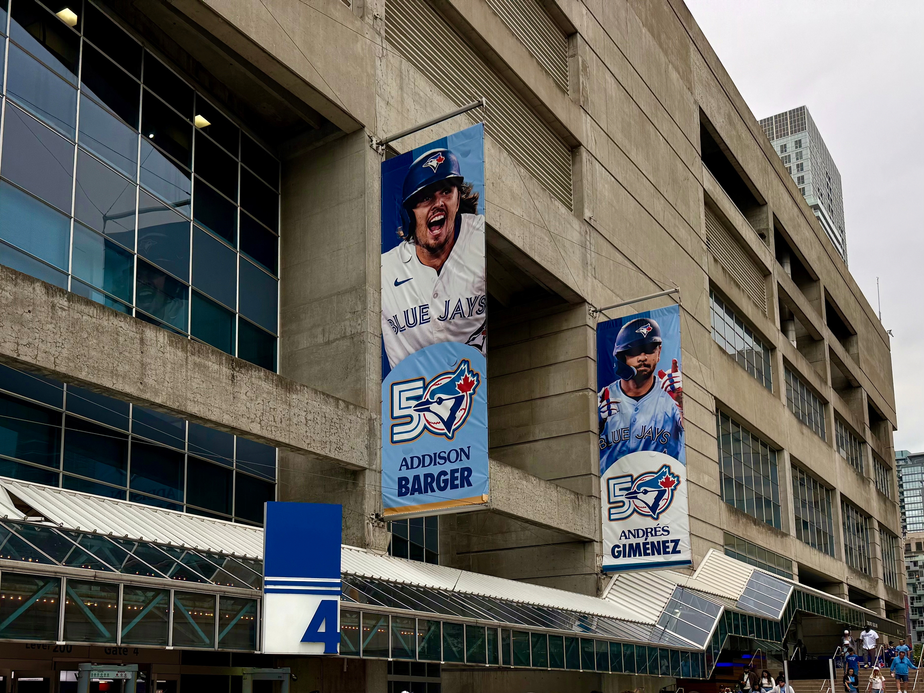The photo shows the exterior of a large concrete stadium with multiple rows of windows and a glass canopy over the entrance. Two tall vertical banners hang on the wall. The left banner features a baseball player in a Blue Jays uniform, wearing a helmet with his mouth open as if shouting, next to the Blue Jays 50th anniversary logo and the name “ADDISON BARGER.” The right banner shows another Blue Jays player wearing a helmet and giving a thumbs-up, with the same 50th anniversary logo and the name “ANDRÉS GIMENEZ.” Below the banners, there is a blue sign with a white number 4 above the entrance. Several people in blue clothing are walking on the pathway near the entrance. A tall modern building is visible in the background to the right under a cloudy sky.