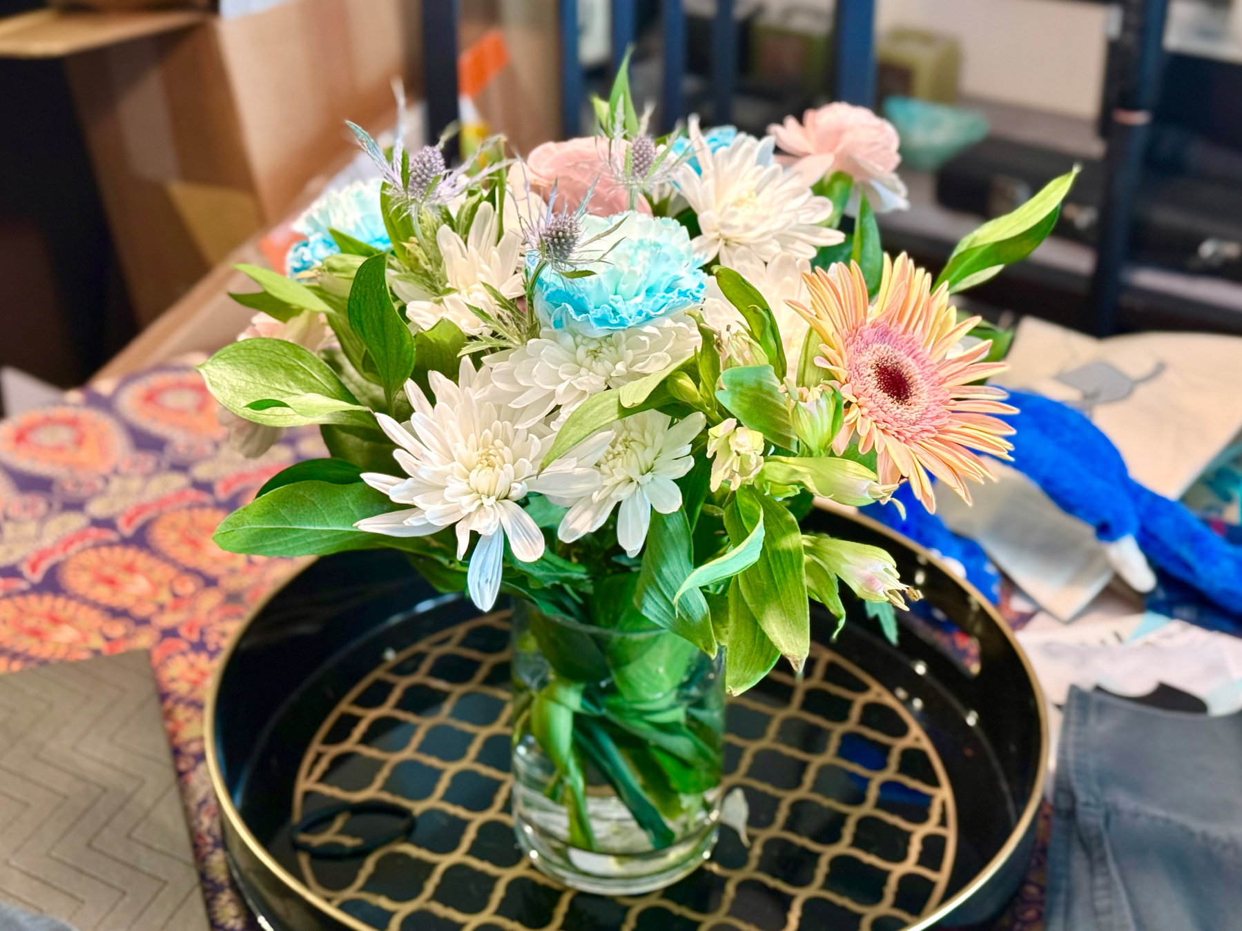 A glass vase filled with a colourful bouquet of flowers sits on a black and gold patterned round tray. The bouquet includes white chrysanthemums, a peach-pink gerbera daisy, pink and blue carnations, and spiky thistle-like flowers, surrounded by lush green leaves. The background shows a patterned tablecloth with red and orange designs, a folded grey napkin, a blue plush toy, and some papers or fabrics. Cardboard boxes and shelving are visible in the blurred background.