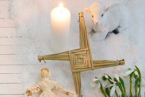 A scene depicting various traditional symbols: a woven straw cross lies on a snowy surface next to a lit white candle. Nearby, a small lamb figurine and a doll made of straw are positioned together. Snowdrop flowers are arranged at the bottom right corner, adding a touch of greenery to the wintry setting.