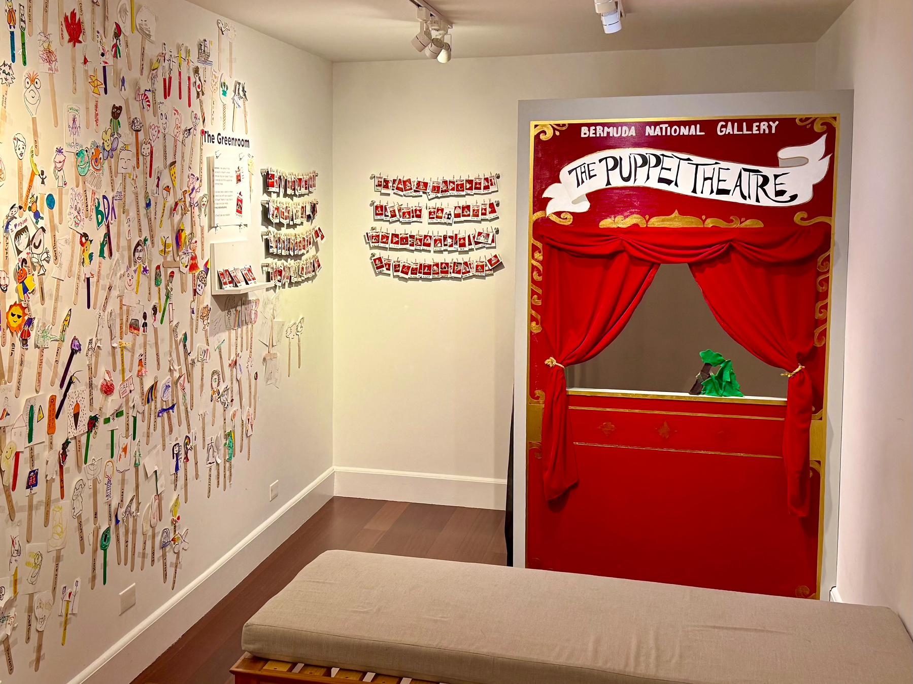 A small, brightly lit room in a gallery features a children’s puppet theatre setup. On the right side, there is a red puppet theatre with gold detailing, red curtains tied to the sides, and a small opening where a green puppet is visible. Above the theatre, the sign reads “Bermuda National Gallery” and “The Puppet Theatre.” On the left side, the walls are covered with numerous handmade paper puppets attached to wooden sticks, decorated with colourful drawings of faces, animals, and patterns. In the centre of the left wall, a sign titled “The Greenroom” is mounted, surrounded by additional small photos and Polaroid-style images. A light beige cushioned bench is placed in the middle of the wooden floor, facing the puppet theatre. The space has a creative and playful atmosphere, designed for children’s interaction.