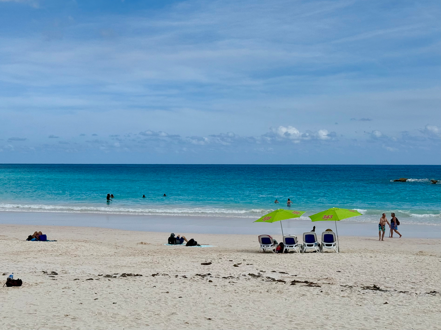 A sandy beach with light beige sand stretches out to a turquoise ocean under a partly cloudy blue sky. Several people are seen in the water enjoying the waves, while a few others relax on the sand closer to the shore. In the centre-right of the image, four white lounge chairs with green umbrellas are set up, and the umbrellas display the number “343” in red. A couple is walking along the shoreline, and small patches of seaweed are scattered on the sand. In the far right of the ocean, a small rocky outcrop is visible.