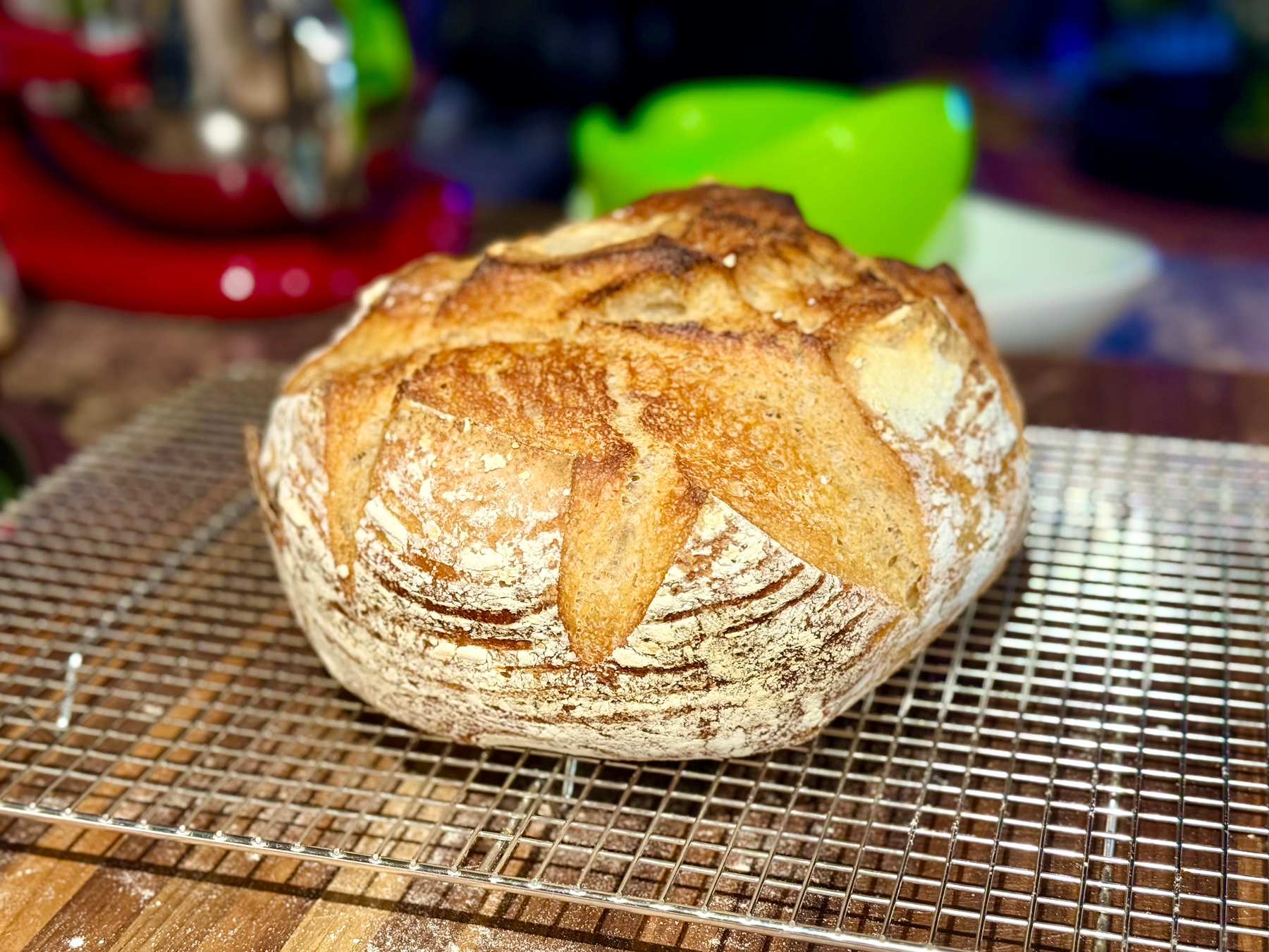 A freshly baked round loaf of artisan bread with a golden-brown crust and star-shaped scoring rests on a metal cooling rack. The crust shows a mix of crispy textures and lightly floured areas, with deep ridges from the scoring pattern. In the background, there is a blurred kitchen scene with a red stand mixer, a green object, and a white plate. The surface beneath the rack is a wooden countertop dusted lightly with flour.