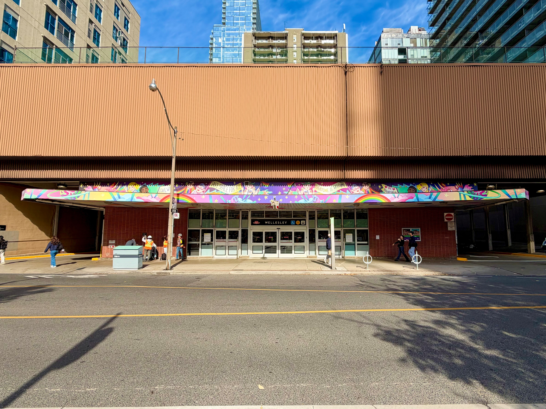 A city street scene shows the entrance to a building with a wide overhang decorated with a colourful mural featuring rainbows, stars, and diverse cartoon-style faces. The mural includes phrases such as “Love,” “We are here for you,” and “Welcome.” Below the mural, the entrance consists of multiple glass doors with a sign above that reads “Wellesley.” Several people are visible outside—some walking, some standing, and a few wearing construction vests. There are tall residential buildings in the background, partially obscured by the brown, ribbed facade of the structure. The street in front is empty, and shadows from trees fall across the pavement.