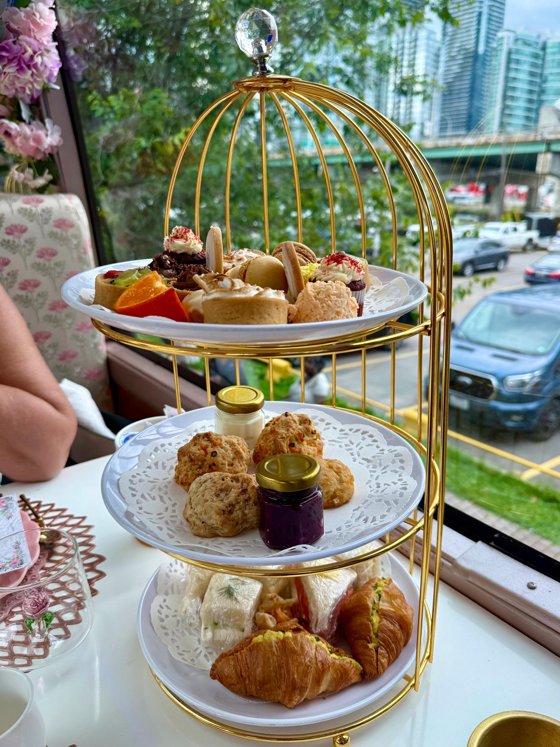 A three-tiered golden birdcage-style stand holds an assortment of afternoon tea treats by a window with a view of cars parked outside and tall city buildings in the background. The top tier features an array of colourful desserts, including macarons, fruit tarts, cupcakes with cream and sprinkles, and small cakes. The middle tier holds several scones, accompanied by small jars of jam and cream. The bottom tier has savoury items, including croissants filled with egg salad and small triangular finger sandwiches wrapped in white bread. A floral teacup and glass teacup with a pink rose design sit on the white table beside the stand, along with a decorative pink lattice placemat. A person with a bare arm is partially visible on the left, and the seating features a floral-patterned chair.