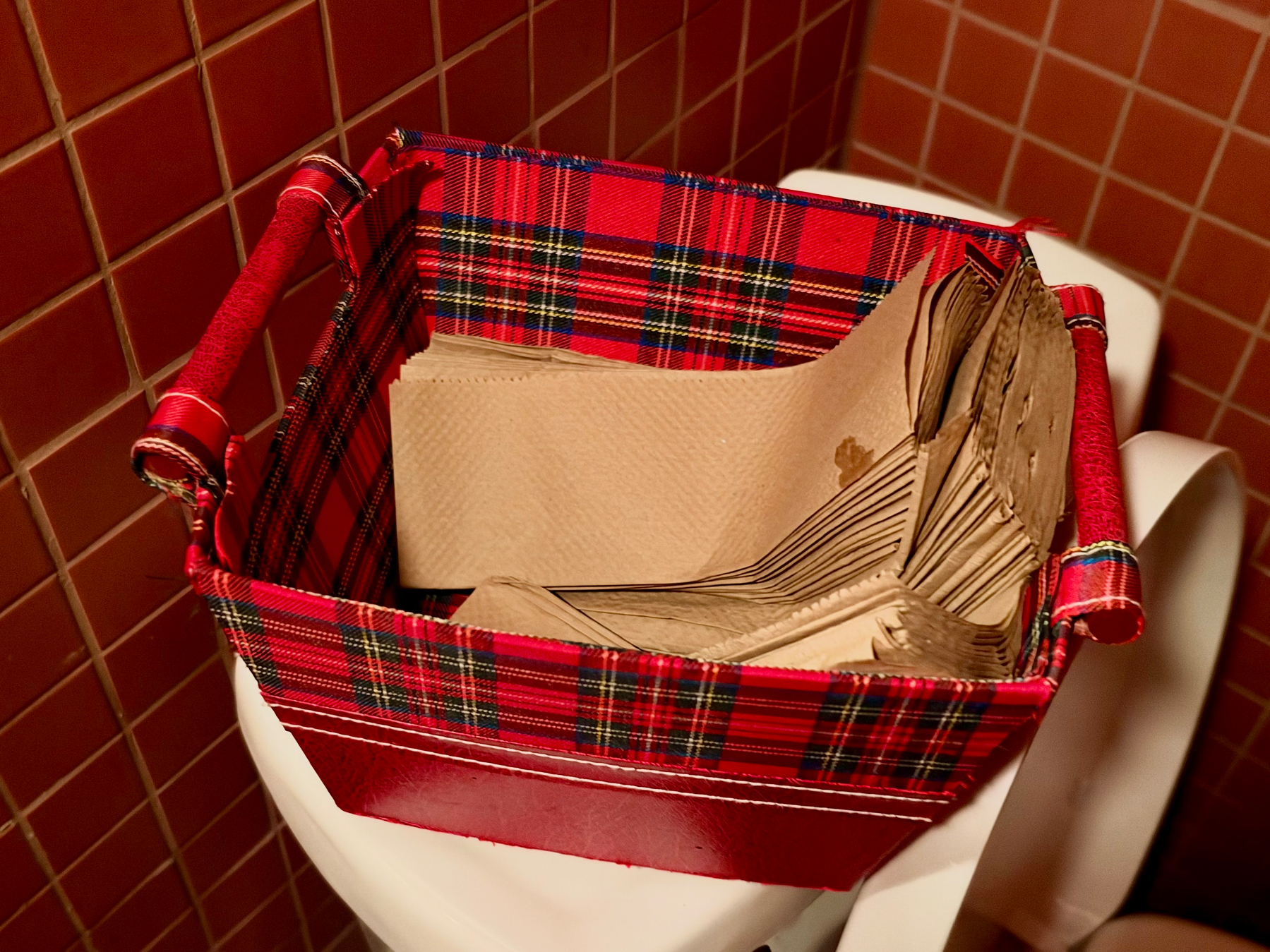 A red and tartan-patterned basket sits on top of a white toilet tank in a bathroom with reddish-brown tiled walls. The basket contains a stack of brown paper napkins or paper towels, some of which appear slightly crumpled. The basket has red handles wrapped in shiny material, and the tartan lining features a mix of red, black, white, and yellow stripes. Another white toilet tank is partially visible beside it.