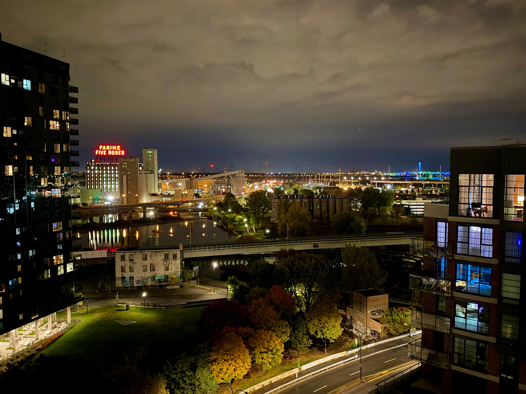 A nighttime cityscape shows a view over a river with lights reflecting on the water. On the left, a tall dark apartment building with many illuminated windows stands next to a brightly lit red sign reading “FARINE FIVE ROSES” atop an industrial building complex. The river runs through the middle of the image, bordered by green spaces and trees with autumn foliage in shades of orange, yellow, and green. Bridges cross the river, and graffiti-covered structures can be seen near the roads. On the right, a modern glass-fronted building with balconies reveals warmly lit interiors. In the distance, an illuminated blue bridge and scattered city lights brighten the horizon under a cloudy night sky.