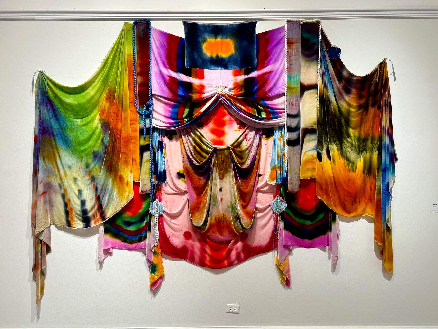 A vibrant textile art installation hangs on a white wall, composed of multiple draped and layered fabrics in a range of vivid colours. The fabrics display a tie-dye or hand-dyed effect with gradients and patterns in bright green, orange, yellow, red, blue, pink, purple, and black. The central fabric is pink with symmetrical folds, topped by a multicoloured drape with a rainbow-like pattern, and flanked by two large side panels in green-orange and yellow-brown blends. Long strips of fabric hang down in various lengths, some with soft, fluid folds, creating a cascading effect. A small section of light blue rope or cord decoratively ties part of the fabric on the left. The installation has an overall flowing, theatrical appearance, resembling a colourful abstract curtain.