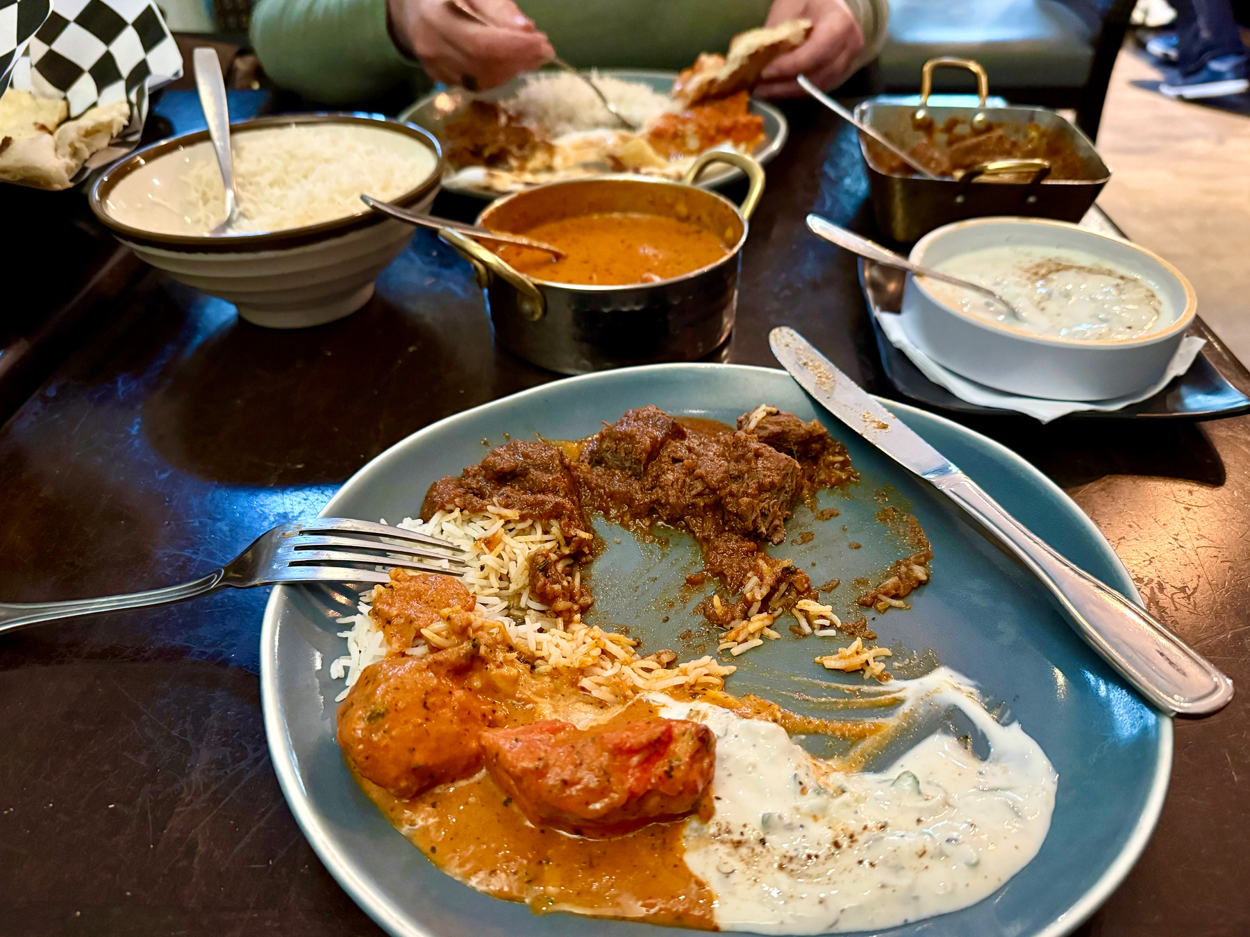 A dining table with an Indian meal partially eaten. In the foreground, a blue plate has basmati rice, pieces of chicken in orange curry, tender chunks of beef or lamb in brown curry, and a portion of white yogurt sauce with herbs. A fork and knife rest on the plate. On the table, there is a metal pot of orange curry, a white bowl of plain rice, a basket of naan bread lined with black-and-white checkered paper, and a white bowl of raita on a black tray. Across the table, another diner in a green sweater is scooping food onto their plate with naan while a rectangular brass dish of another curry sits nearby. The table is dark wood and the restaurant environment is softly lit.