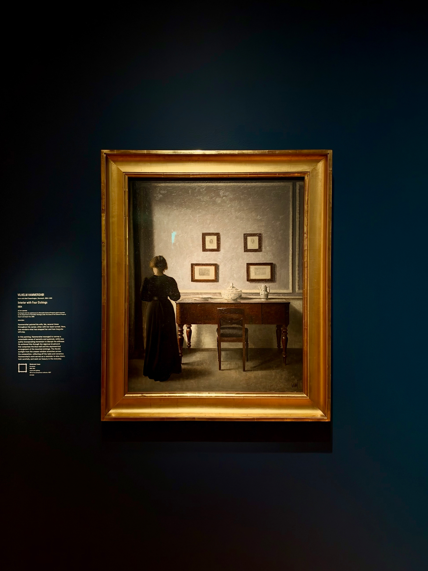 A framed painting hangs on a dark blue wall in a gallery. The painting depicts a woman in a long black dress standing in front of a wooden table with turned legs. She is facing away from the viewer, looking towards a light gray wall where four small framed etchings are evenly arranged in two rows. On the table, there is a white pitcher, a glass, and some papers. A single wooden chair is placed in front of the table, slightly off-center. To the left of the painting, a wall label provides information about the artwork in small white text. The frame is golden and ornately simple, contrasting with the dark wall behind it.