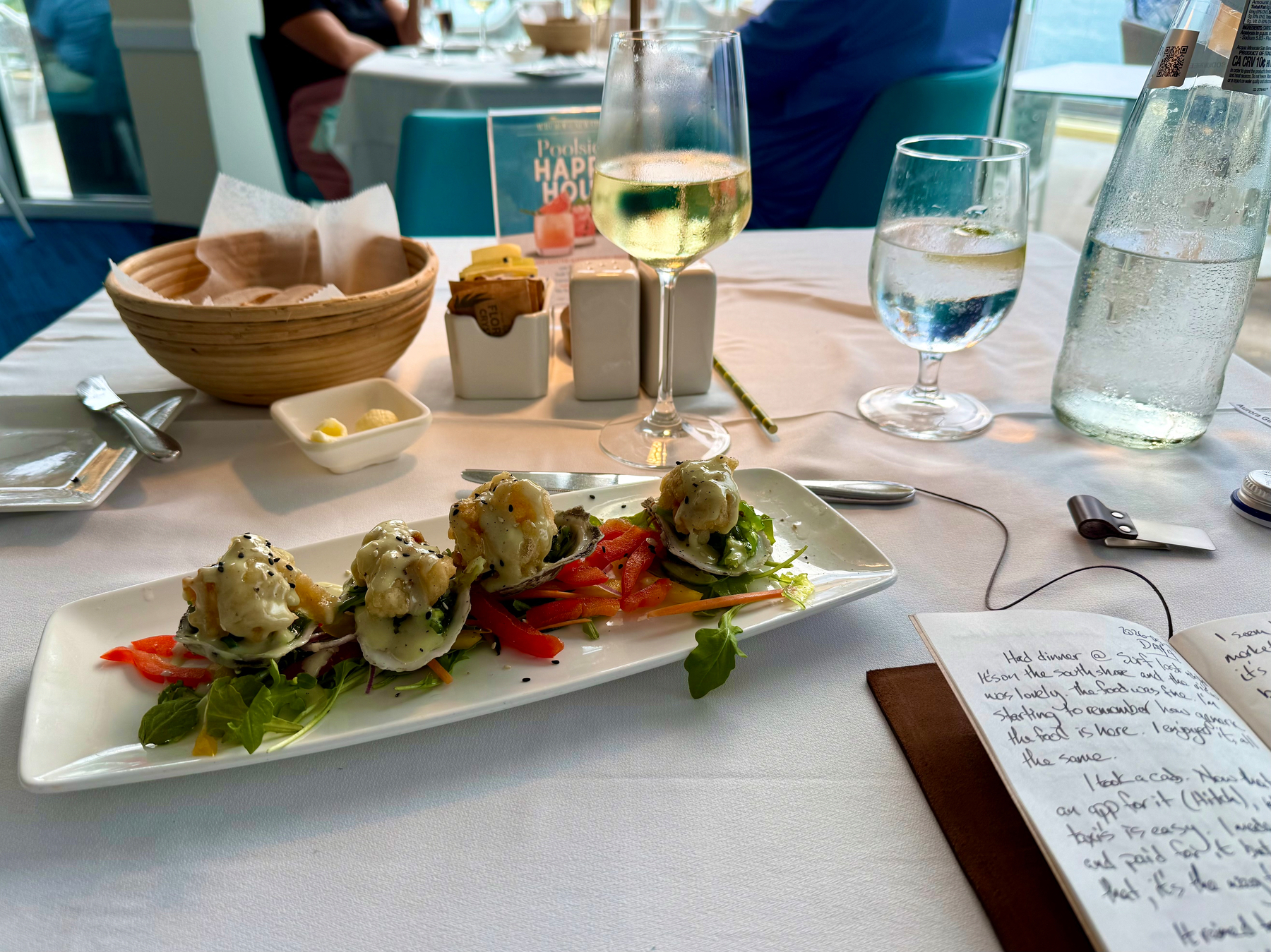A white table at a seaside restaurant is set with a glass of white wine, a glass of water with a lemon slice, and a tall bottle of sparkling water. On the table is a rectangular white plate with four pieces of seafood topped with a creamy sauce and garnished with arugula, shredded vegetables, and sesame seeds. Behind the dish is a basket of bread with a small dish of butter, along with sugar packets and butter portions in a white holder. A handwritten journal lies open on the table, with pages partially filled in black ink. In the background, other tables with white linens and diners are visible, along with a bright window looking out toward the sea.