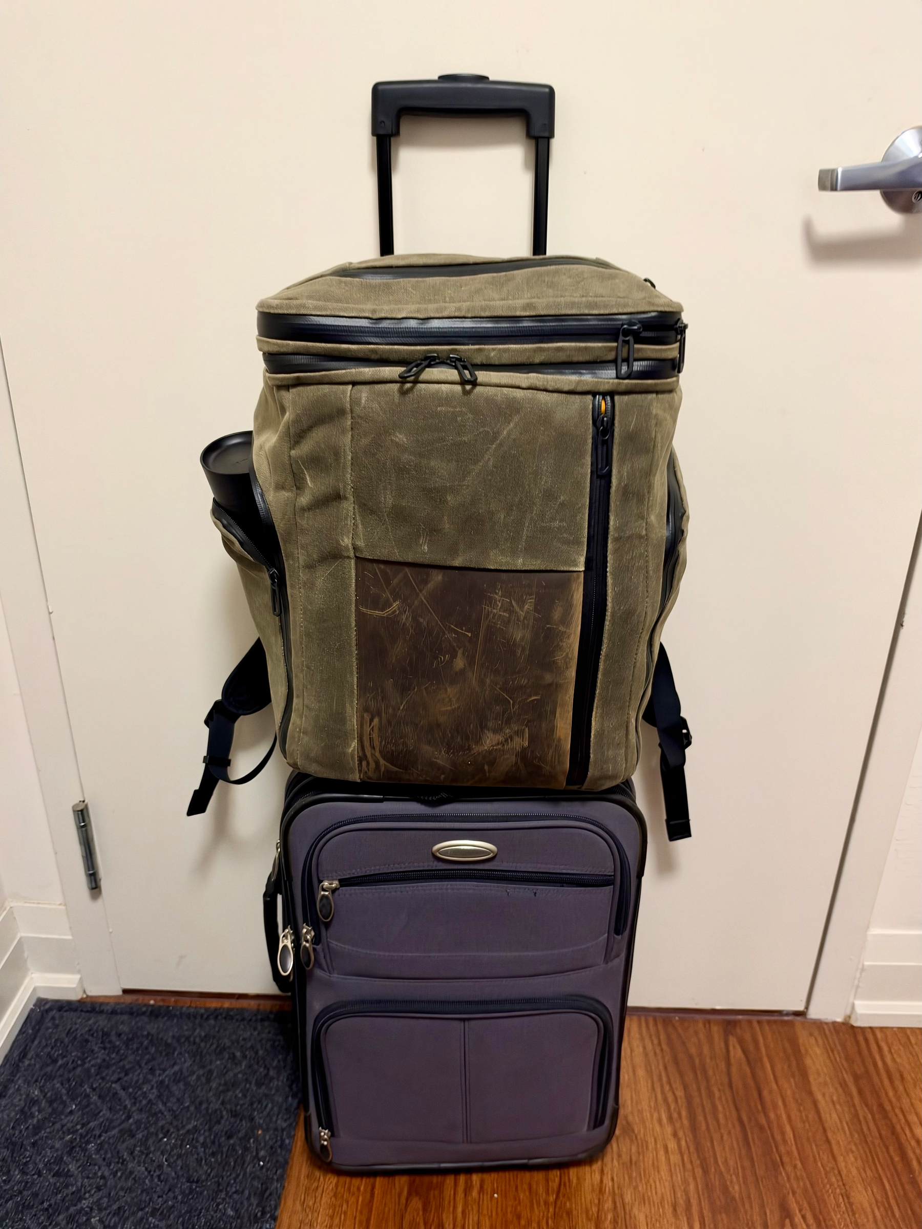 A small rolling suitcase with a telescopic handle extended is placed against a white door with a silver handle. On top of the suitcase is an olive-green, weathered backpack with black zippers and straps. The backpack has a rugged and scratched brown leather-like front panel and a water bottle in the left side pocket. The rolling suitcase is dark grey with a front zippered pocket, two smaller zippers near the top, and a small oval-shaped silver emblem. The floor is a combination of wooden panels and a dark textured mat in front of the door.