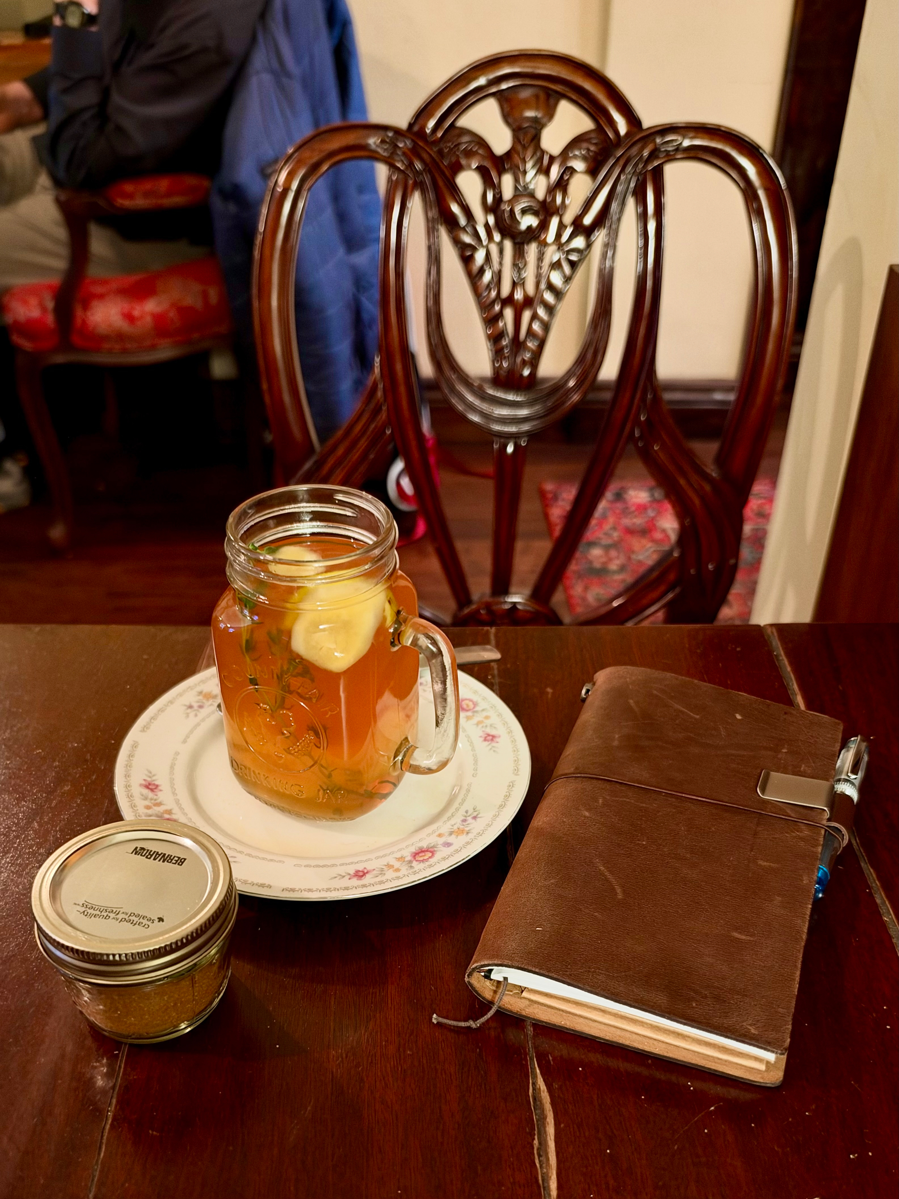 A wooden table in a cozy café with several items on it. On the left is a small glass jar with a golden lid, filled with a yellowish-brown granular substance. In the center is a mason jar mug with a handle, filled with a light amber drink garnished with a lemon slice and sprigs of herbs, placed on a white floral-patterned saucer. To the right is a brown leather-bound notebook with a pen clipped under its elastic strap, slightly open with pages visible. In the background is a dark wooden chair with an ornate carved backrest, and another table with red-upholstered chairs, one holding a person wearing khaki pants, a dark sweater, and a blue jacket draped over the back. The floor is wooden with a red patterned rug partly visible.