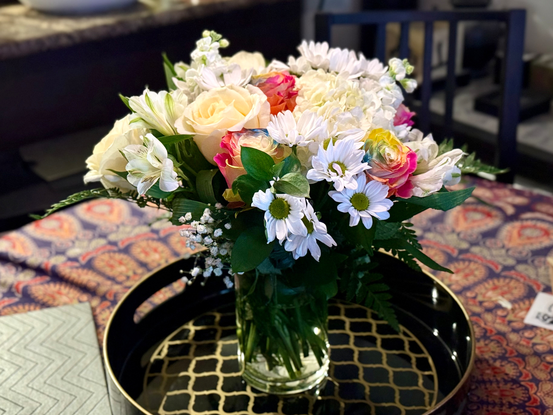 A bouquet of fresh flowers in a clear glass vase filled with water, placed on a black and gold patterned tray. The bouquet includes cream and pink roses, white daisies with yellow centres, white alstroemeria, small white filler flowers, and green leaves. The table beneath the tray is covered with a patterned fabric featuring red, orange, and blue designs. In the background, a dark wooden chair and part of a kitchen counter are visible, slightly out of focus.