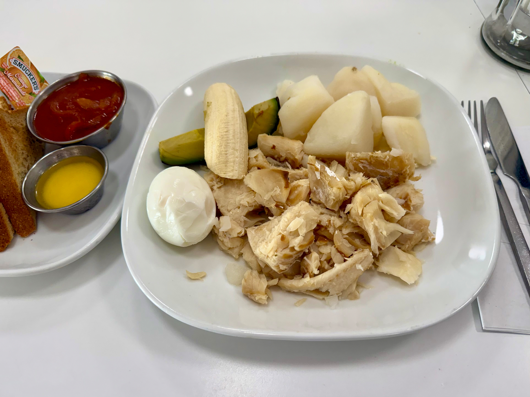 A white plate with a meal including shredded salted codfish, a peeled boiled green banana, a boiled egg, boiled pieces of yam, and a piece of peeled boiled plantain. To the left, there is a smaller plate with toast slices, a foil packet of Smucker’s orange marmalade, a small metal container of melted butter, and another metal container of tomato-based sauce. A fork and knife rest on a napkin to the right of the main plate, and the setting is on a white tabletop.