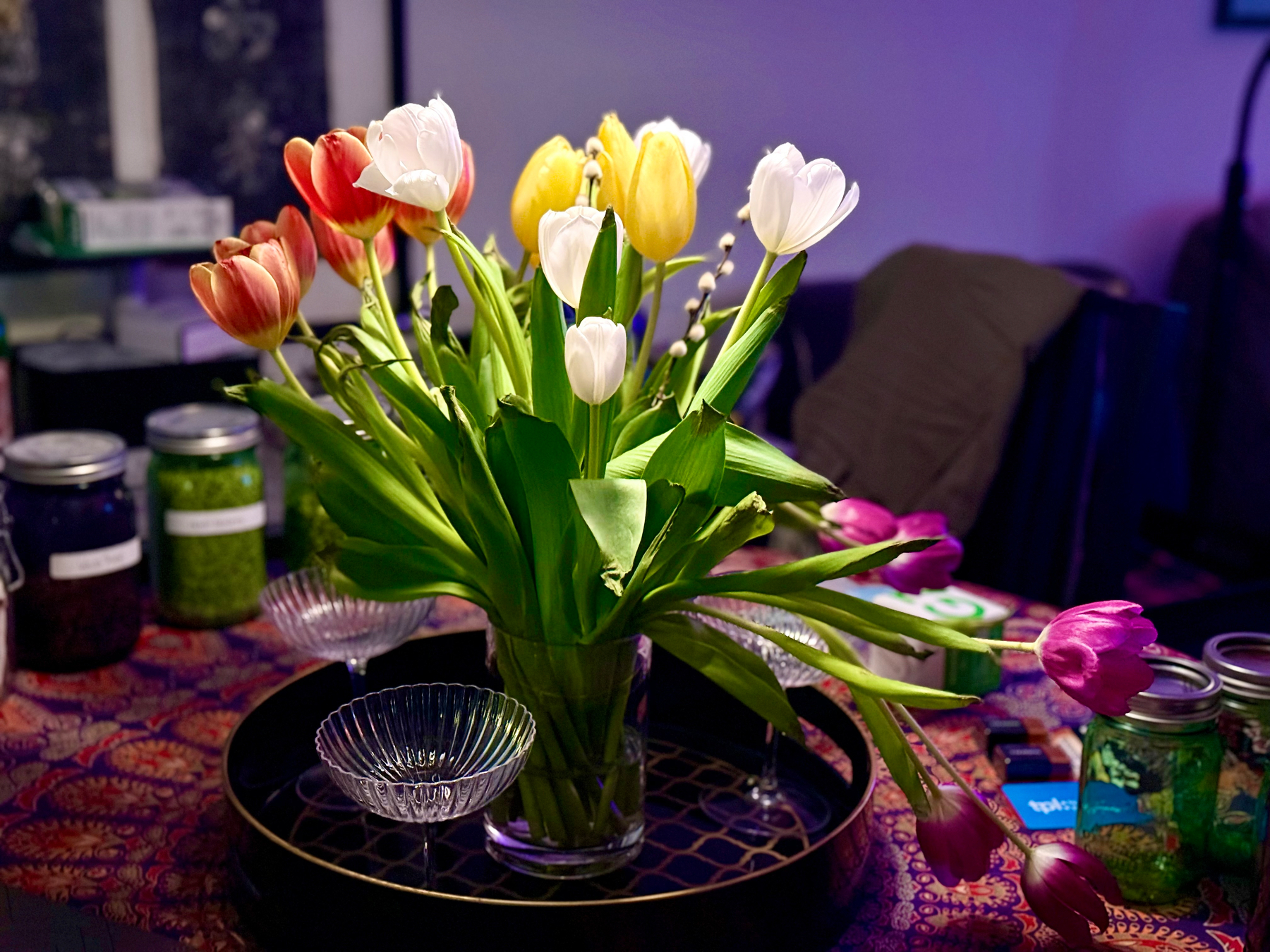 A bouquet of tulips in various colors, including red, white, yellow, and purple, is arranged in a clear vase on a patterned tablecloth. The table also holds a few glass jars, some with green contents, and small dishes on a decorative tray. The background is softly lit, creating a cozy ambiance.