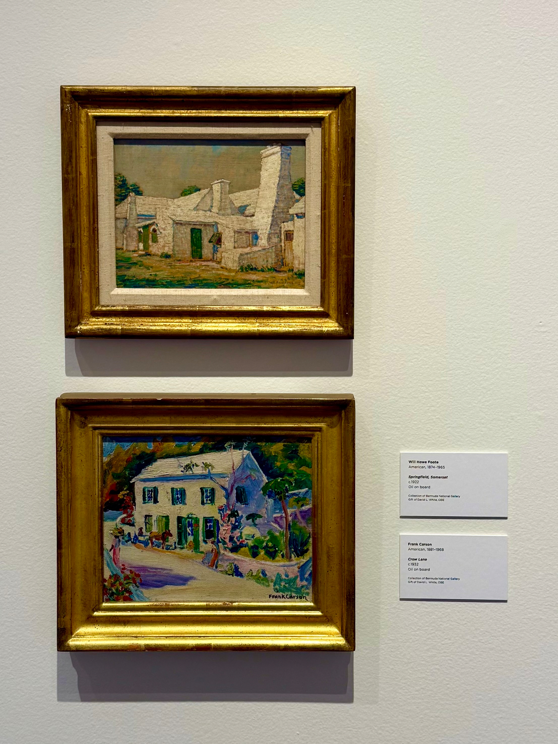 Two framed paintings hang on a white gallery wall, each in a gold frame. The top painting depicts a row of stone cottages with steeply pitched roofs and tall chimneys, with a muted colour palette of greys, greens, and browns. The scene is calm, with a simple foreground of dirt and sparse grass. The bottom painting shows a lively, colourful house with green shutters and surrounded by bright greenery and flowers, with several figures and animals around the yard. This painting is more vibrant, with shades of blue, purple, pink, and green. Beside the paintings are two small rectangular museum labels. The top label identifies the upper painting as “Springtime, Etaples” by Will Howe Foote, an American artist (1874–1965), oil on board. The lower label identifies the bottom painting as “Crecy Lisse” by Frank Carson, an American artist (1891–1968), oil on board.