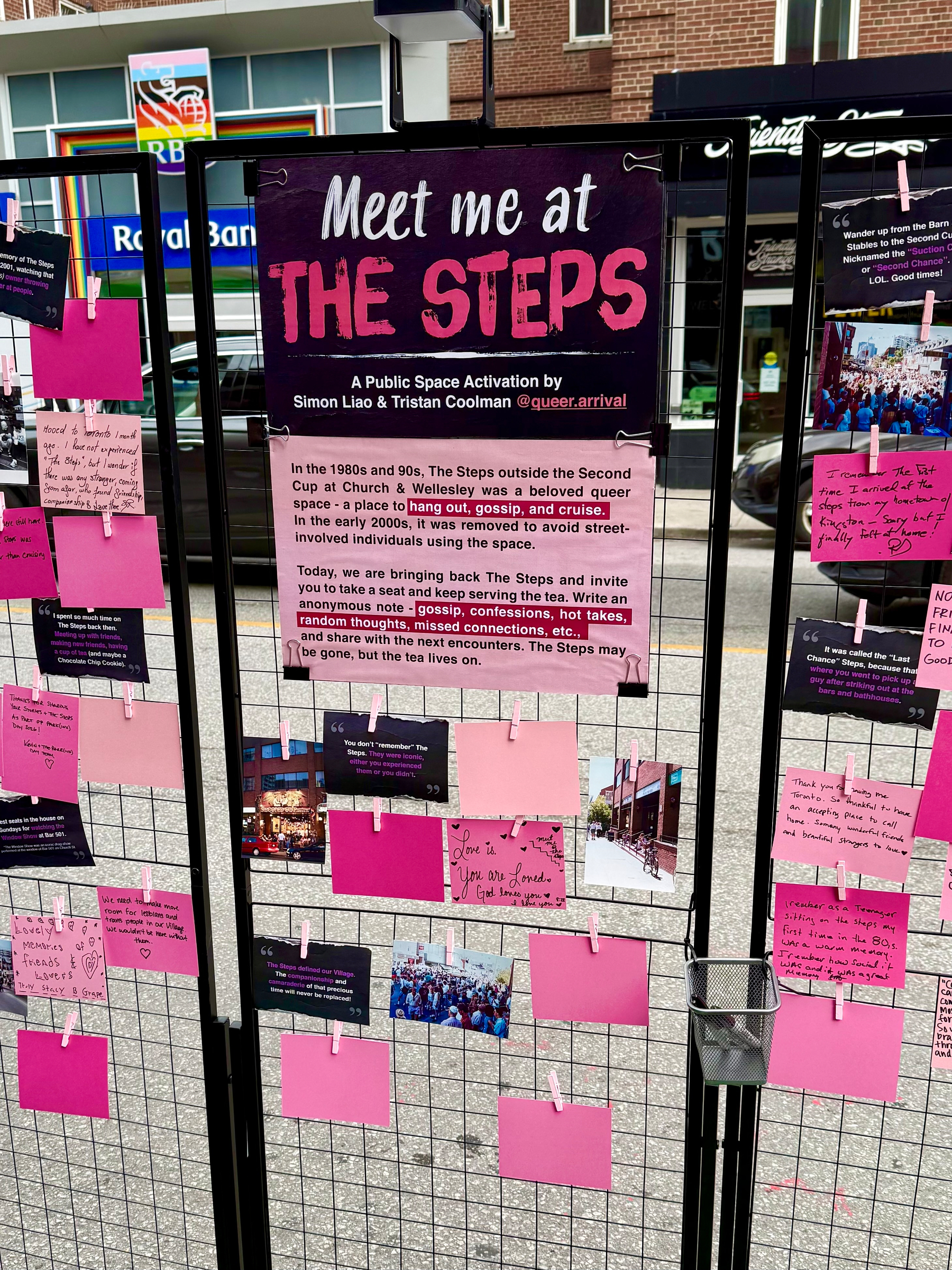 A public art installation is set up on a city sidewalk in front of a Royal Bank branch and other storefronts. The installation is a black metal grid structure with multiple panels displaying pink, purple, and black cards, some with handwritten notes and printed messages. A large central sign reads “Meet me at THE STEPS” with a description below that explains it as a public space activation by Simon Liao and Tristan Coolman, focusing on recreating the queer gathering space once known as The Steps outside the Second Cup at Church and Wellesley. The sign invites people to sit, write anonymous notes such as gossip, confessions, hot takes, and random thoughts, and leave them for others to see. Many pink and purple cards are clipped to the wire panels, some containing handwritten notes and some blank. There are photographs attached, including images of crowds, a street scene with a brick building, and community gatherings. A small metal basket is attached to the display, holding additional cards or supplies. The street and sidewalk around the installation are visible, with cars and parts of buildings in the background.