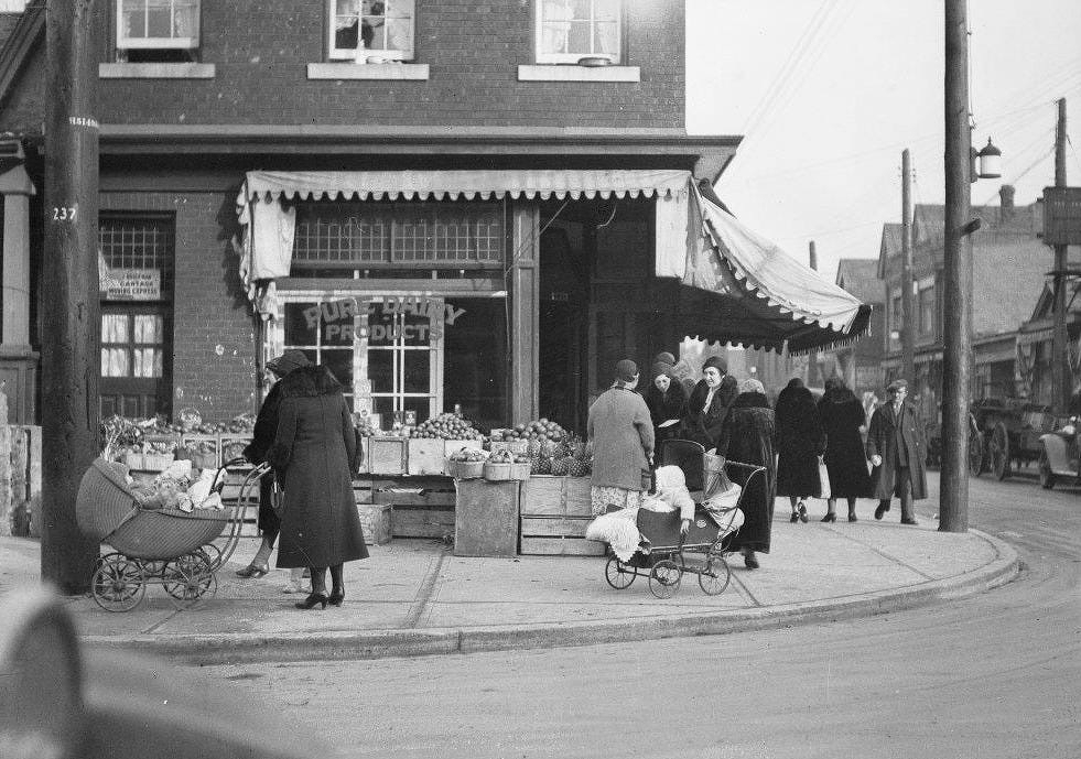A black and white photo of a street scene by a corner store with an awning and sign displaying "Pure Dairy Products." A group of women, dressed in early 20th-century style coats and hats, are gathered in front of the store. Two women are pushing vintage baby carriages with infants inside. Various crates of produce are displayed in front of the store. Several pedestrians are walking along the sidewalk, and a few cars and buildings are visible in the background.