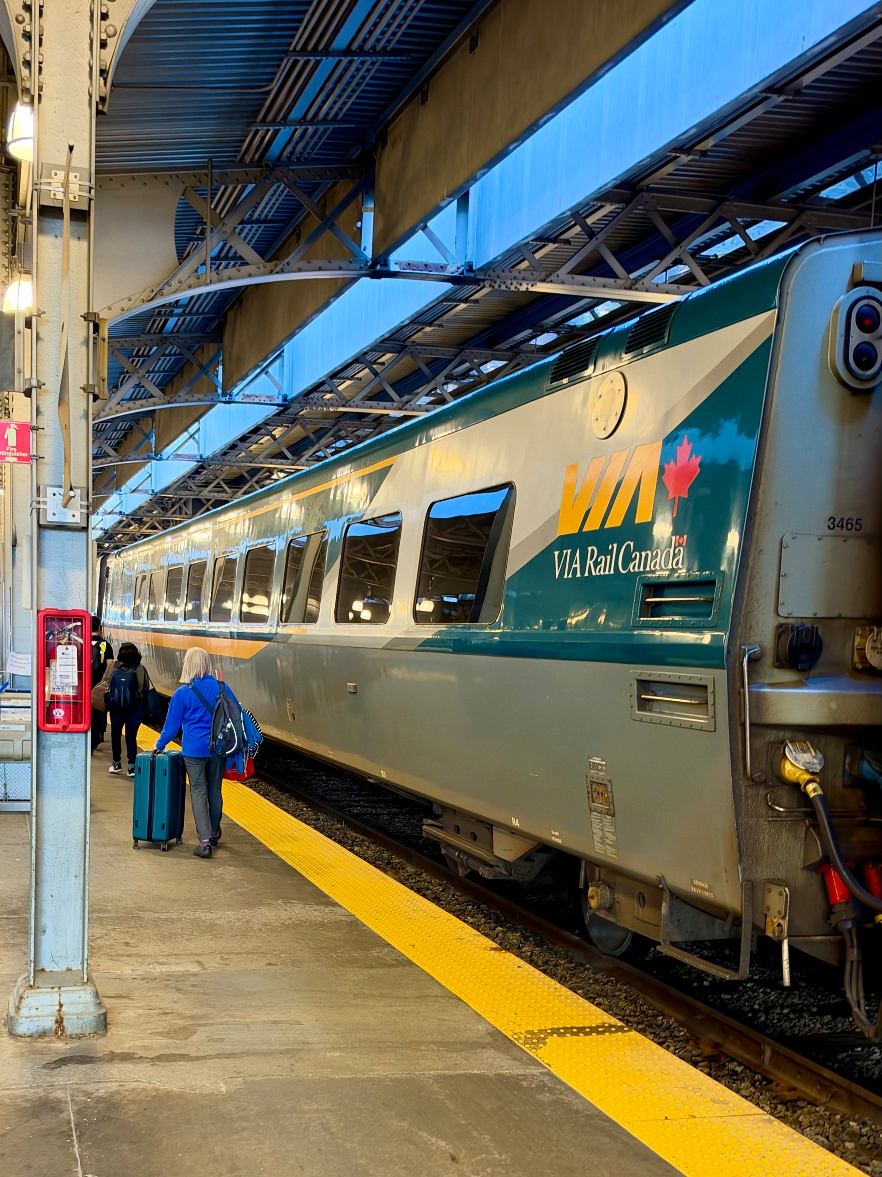 A VIA Rail Canada train is stationed at a covered platform. The train is silver with a teal and yellow stripe, and the VIA Rail logo features a red maple leaf. Several passengers are walking along the platform, with one person in a blue jacket pulling a teal suitcase and carrying a bag. The platform has a bright yellow tactile strip along the edge, and the area is lit by overhead lights. A red emergency call box is mounted on a steel support column, and a pink sign hangs above indicating the direction to the women’s washroom. The roof structure includes metal beams and panels with openings to allow natural light.