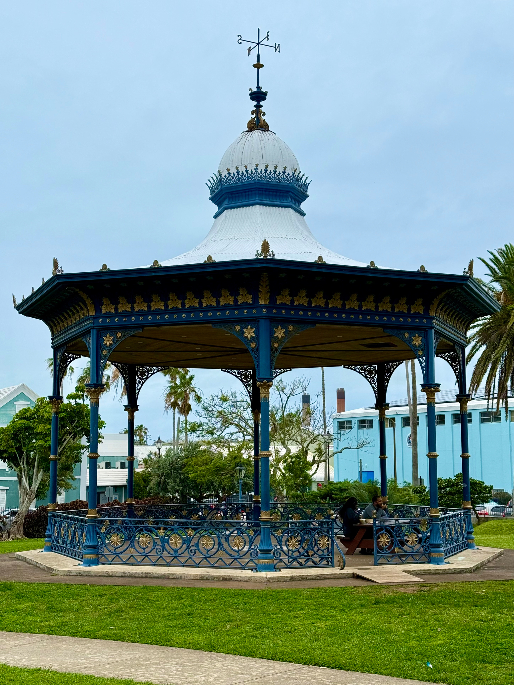 A decorative blue and gold octagonal gazebo with an ornate white domed roof sits in a grassy park. The roof has intricate gold accents and a metal weather vane on top with the cardinal directions N, S, E, and W. The gazebo features wrought-iron railings with star and floral patterns, and several people are seated on benches inside. In the background, there are palm trees, leafy green trees, and pastel-coloured buildings, including a light blue building with a taller industrial-looking structure behind it. The sky is overcast.