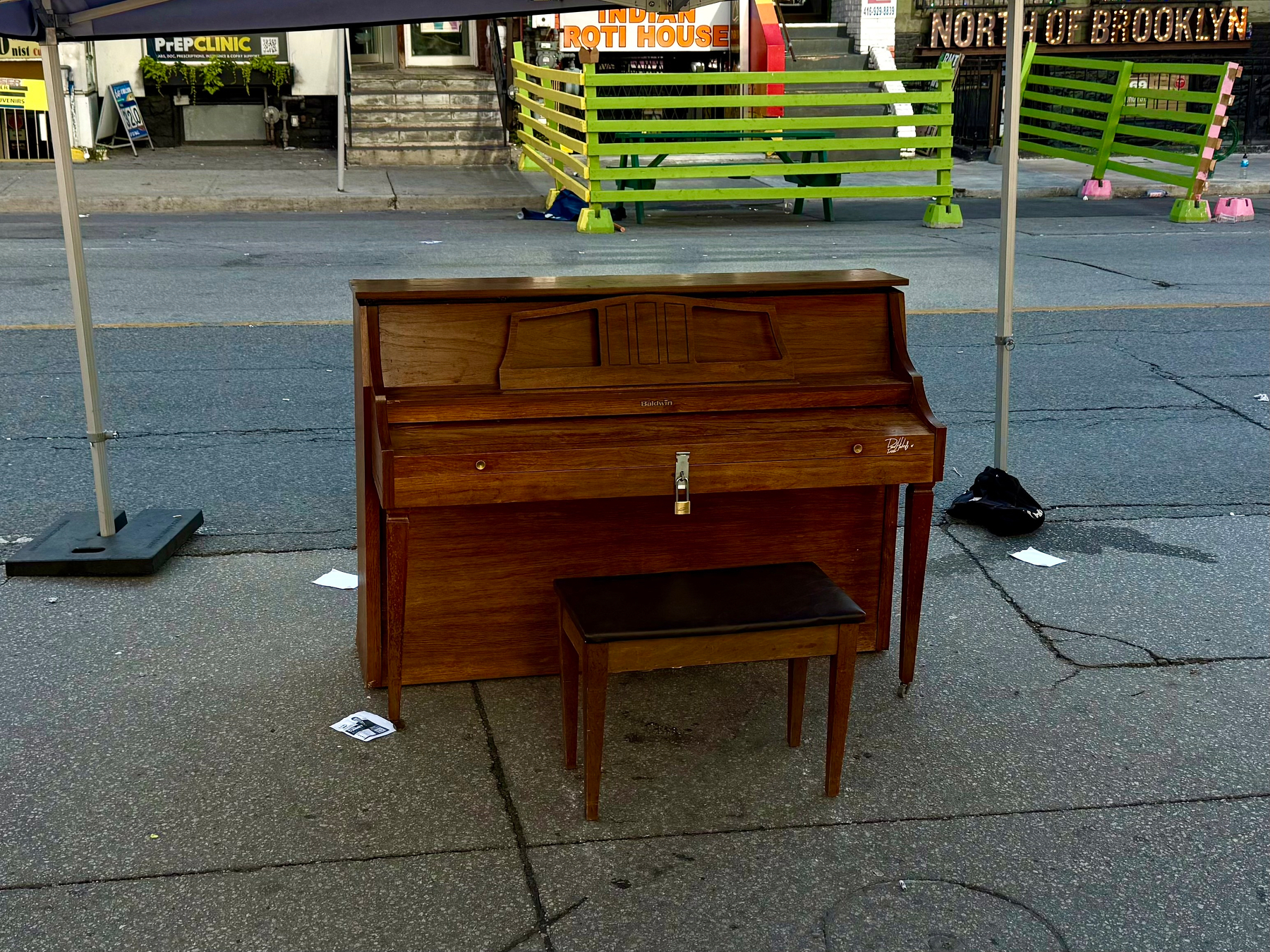 A wooden upright piano with a matching bench is placed on a street under a canopy tent. The piano has a classic design with a small lock on the front panel. Behind the piano, the street is visible with green and pink wooden barriers separating a pedestrian area from the road. In the background, storefronts and signs are visible, including one that says “Indian Roti House” and another that says “North of Brooklyn.”