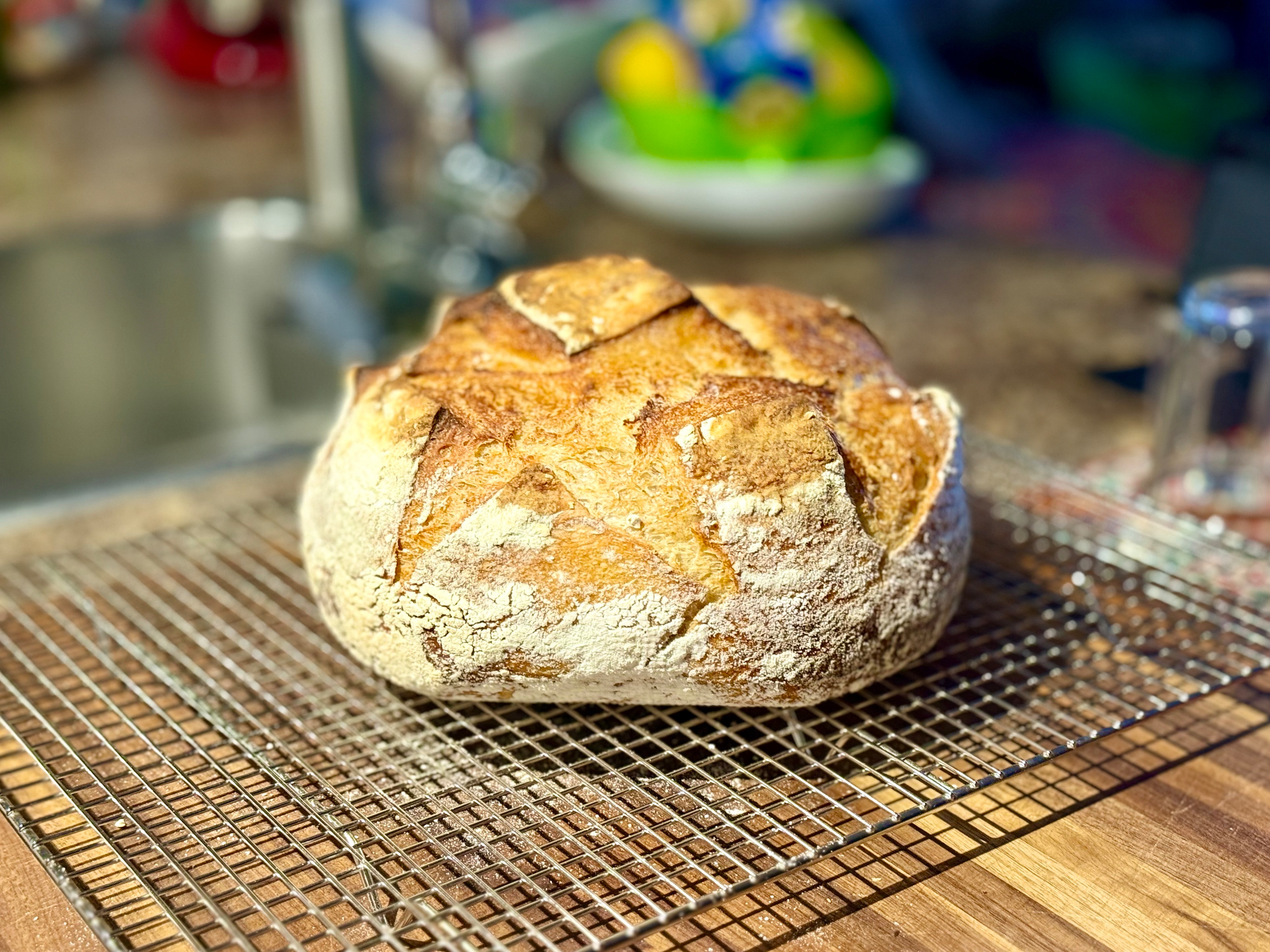 A freshly baked round loaf of rustic bread with a golden-brown crust and deep scoring sits on a metal cooling rack on a wooden countertop. The surface of the bread is lightly dusted with flour, and the crust has a crisp, textured appearance with prominent cracks. In the blurred background, there is a kitchen sink, a faucet, and a bowl of brightly coloured lemons and limes. The overall lighting is warm, highlighting the bread’s crust and texture.