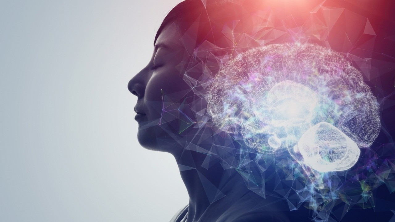 A side profile of a woman with her eyes closed, appearing calm and peaceful, against a light gradient background. Overlaid on her head is a bright, translucent, digital illustration of a human brain, glowing with shades of blue, purple, and white. Abstract polygonal shapes and lines surround the brain, giving a futuristic and technological impression. There are no words or text in the image.