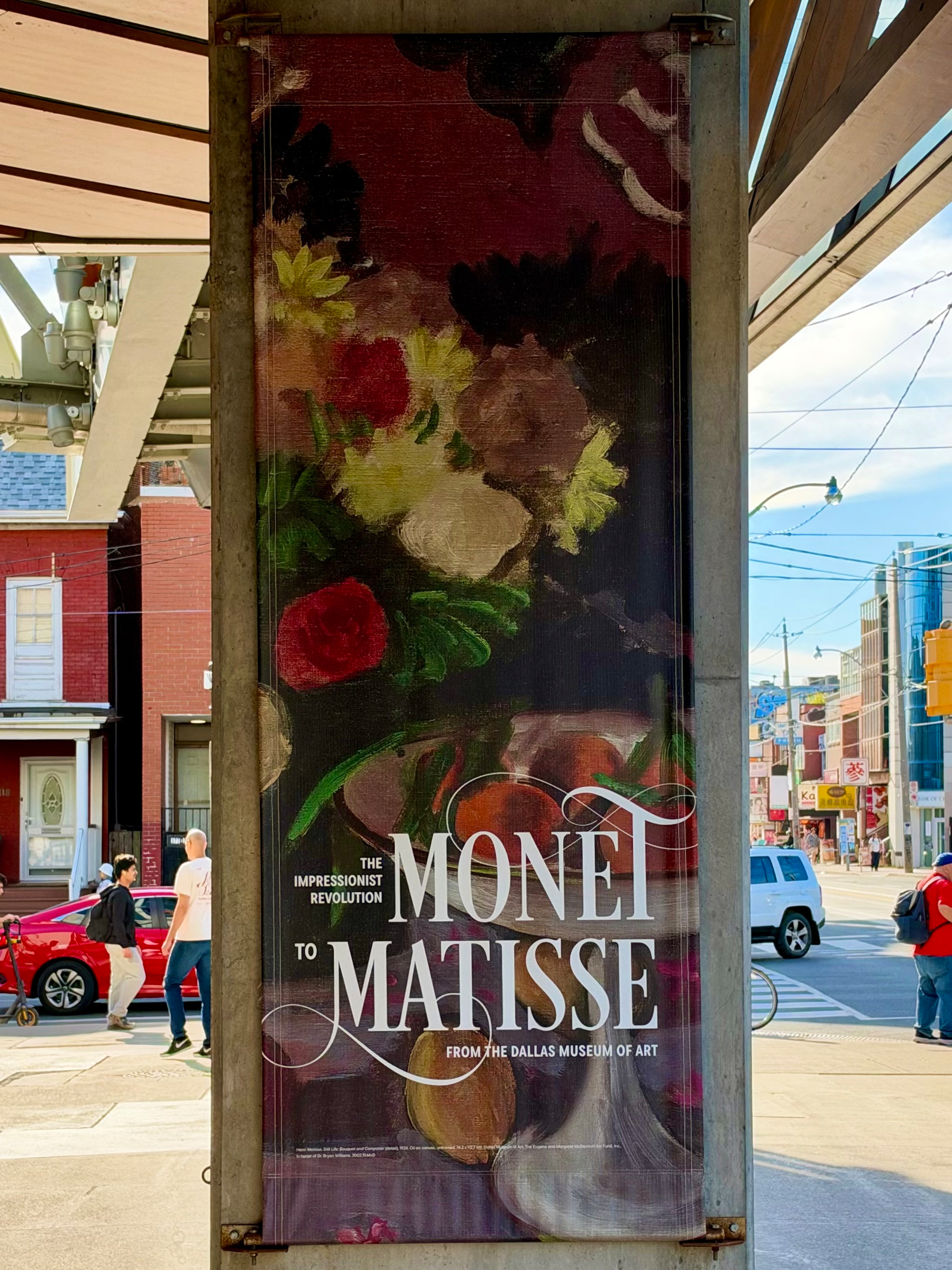 A vertical street banner attached to a metal post displays an art exhibition advertisement featuring an impressionist-style still-life painting of flowers in vivid reds, yellows, greens, and whites against a dark background. The banner text reads: “THE IMPRESSIONIST REVOLUTION MONET TO MATISSE FROM THE DALLAS MUSEUM OF ART” in elegant white lettering with decorative swashes. The banner is located in an urban street setting with a red brick building, residential windows, and a red car in the background. Two men are walking on the sidewalk to the left, and a person with a red backpack is walking to the right across the street. Power lines and a bright sky with some clouds are visible above the buildings.