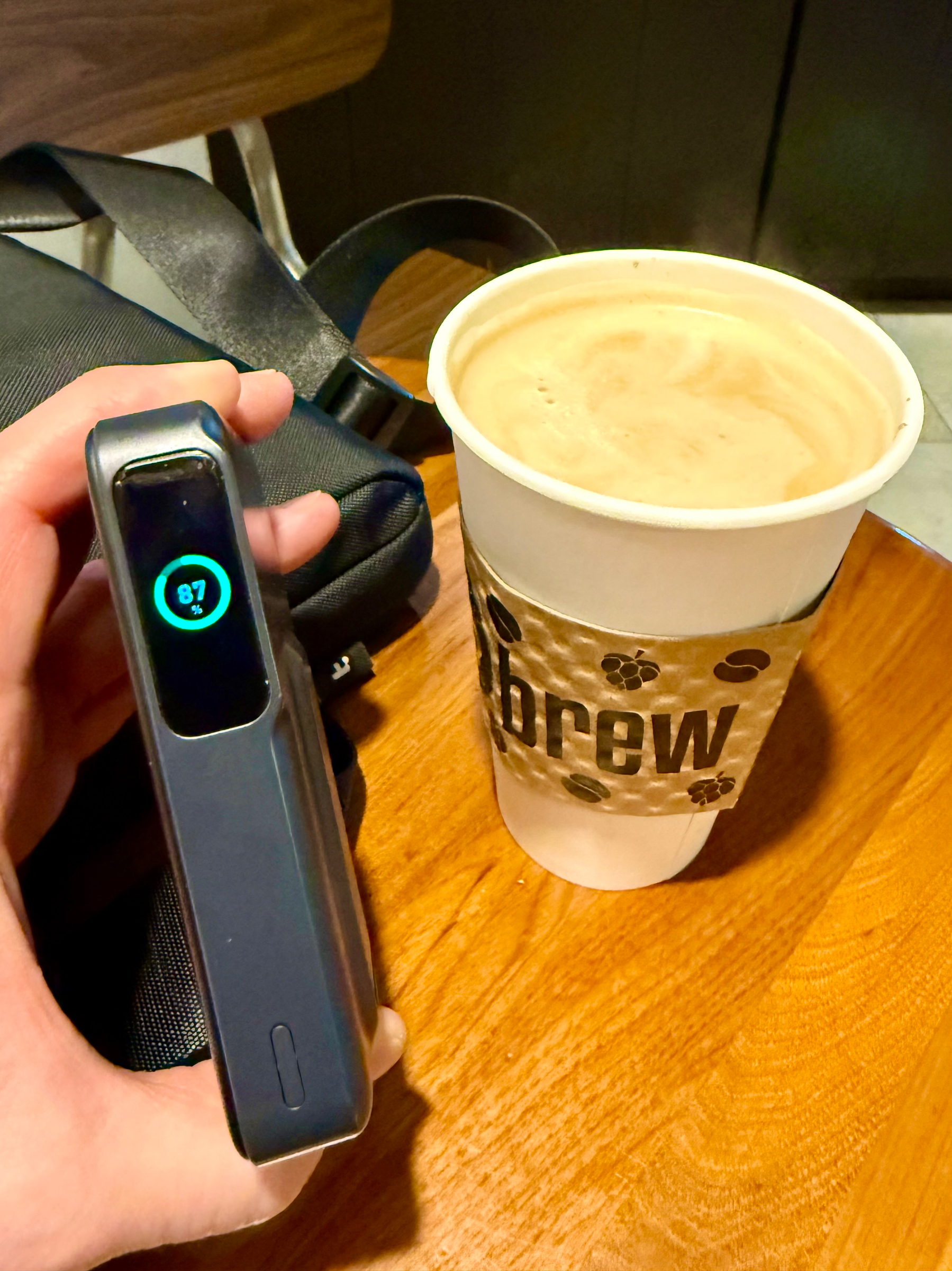 A hand holds a small black electronic device with a screen displaying a green circle and the number “87%.” The device is held over a wooden table next to a white paper cup filled with coffee that has a layer of foam on top. The cup has a textured brown sleeve with the word “brew” and small coffee bean graphics printed on it. In the background, part of a black shoulder bag with a strap is visible on the table, along with a wooden chair and a dark wall.