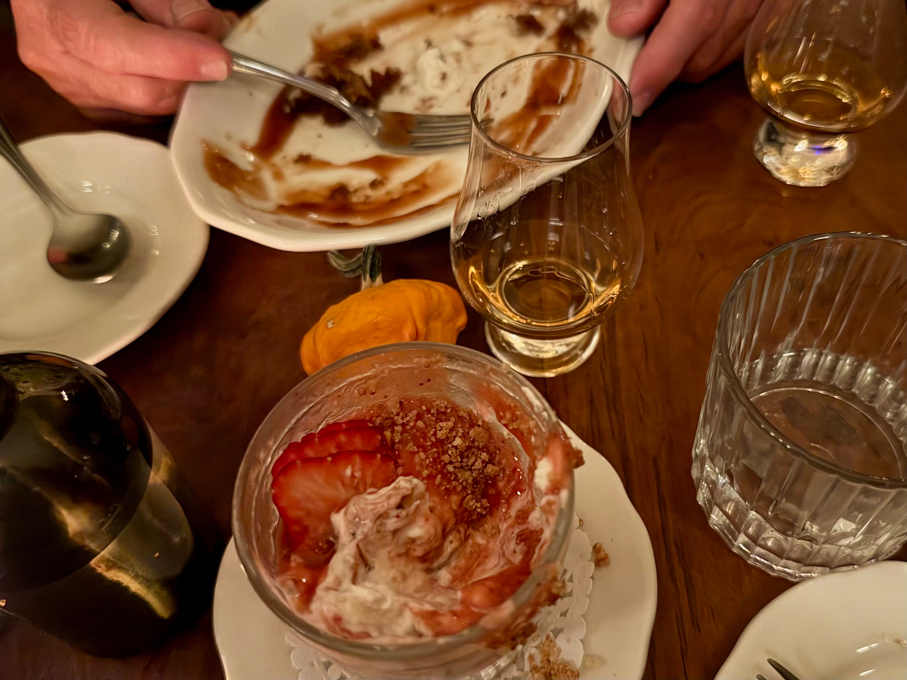 A wooden table is set with various dessert and drink items. In the foreground, there is a glass dessert cup with remnants of whipped cream, strawberry slices, and a crumb topping, sitting on a small white plate with a doily. A mostly empty bottle is visible to the left. In the middle, a small orange gourd is placed next to a glass of amber-coloured liquor. To the right, there is a ribbed glass of water. In the background, a white plate with streaks of caramel or chocolate sauce and bits of dessert is being held and scraped with a fork by a person. Another hand rests on the table near a similar glass with a small amount of amber liquor. A spoon rests in an empty white plate to the left. The scene suggests the end of a shared dessert and drink experience.