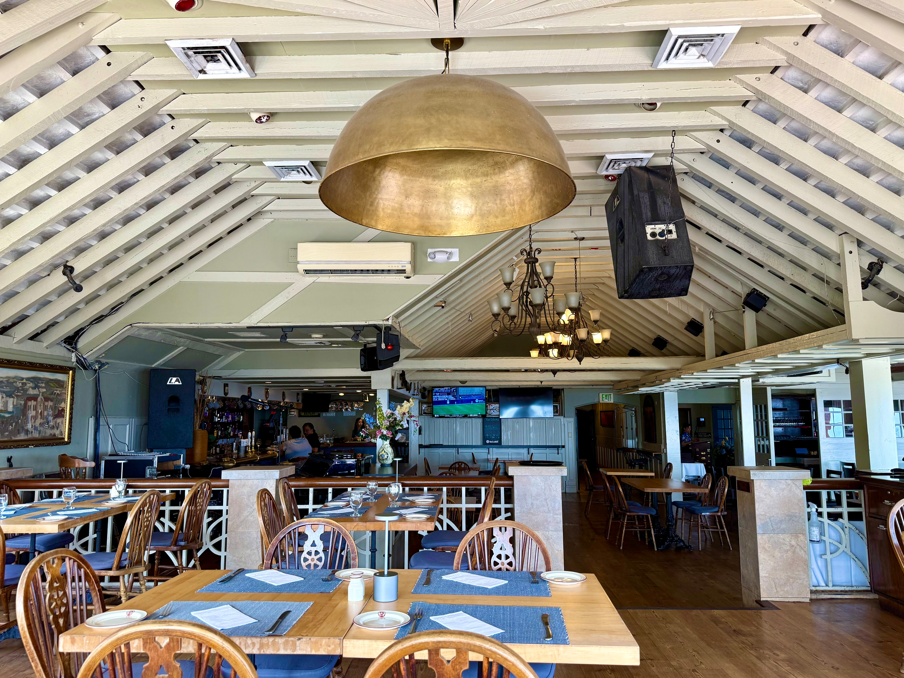 A spacious restaurant interior with wooden tables and chairs arranged neatly, each table set with blue placemats, plates, bowls, glasses, and cutlery. The ceiling is vaulted with exposed light-coloured beams and multiple hanging lights, including a large golden dome pendant and a rustic chandelier. Two black speakers hang from the ceiling, and air conditioning vents are visible. In the background, there is a bar area with a bartender and shelves of bottles, as well as two flat-screen televisions mounted on the wall showing a sports game. The floor is wooden, and the walls are light-coloured with decorative artwork and flowers on a table near the bar.